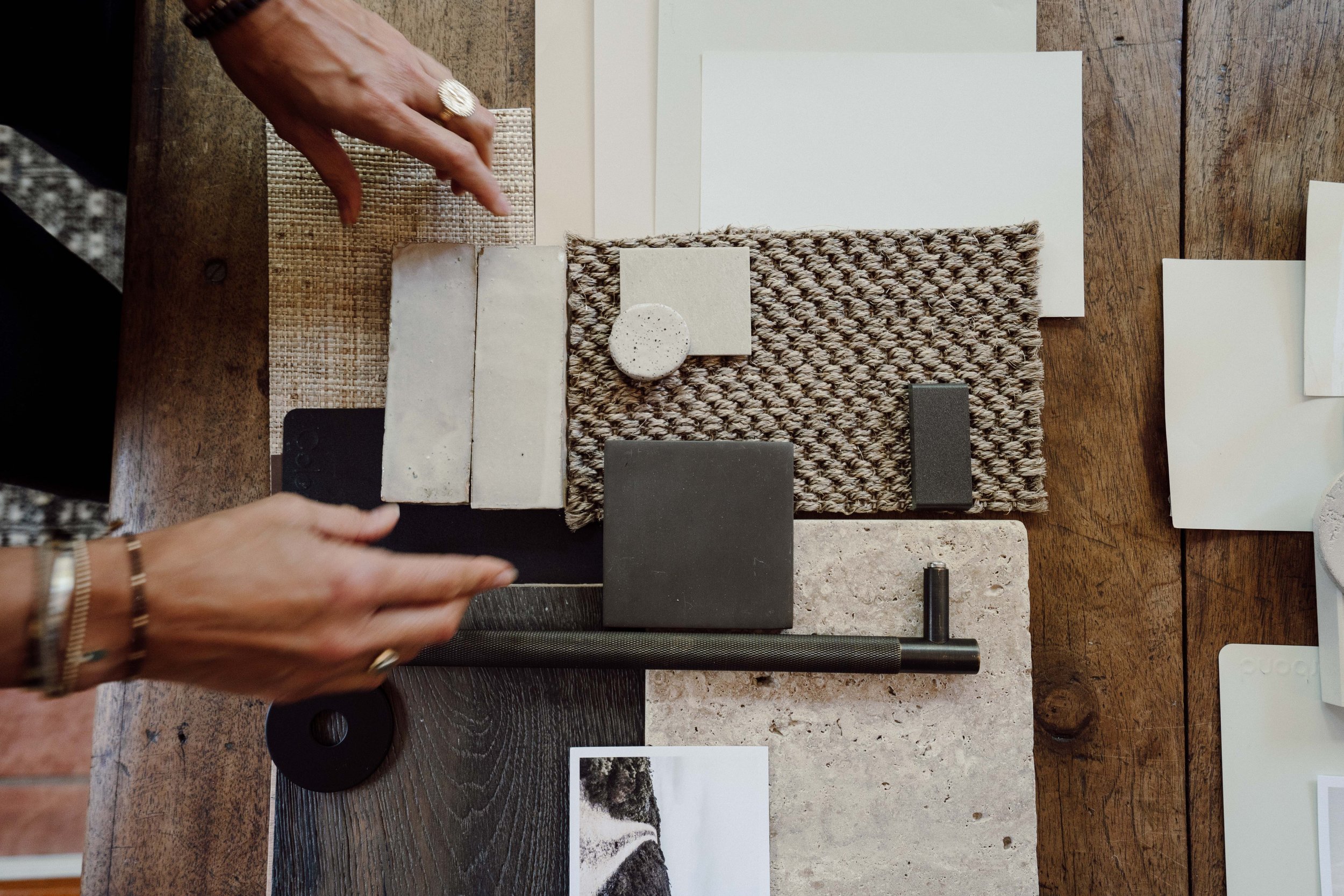 Arrangement of various sample materials on a wooden table, including tiles, fabric swatches, and color samples, with hands pointing towards the samples.