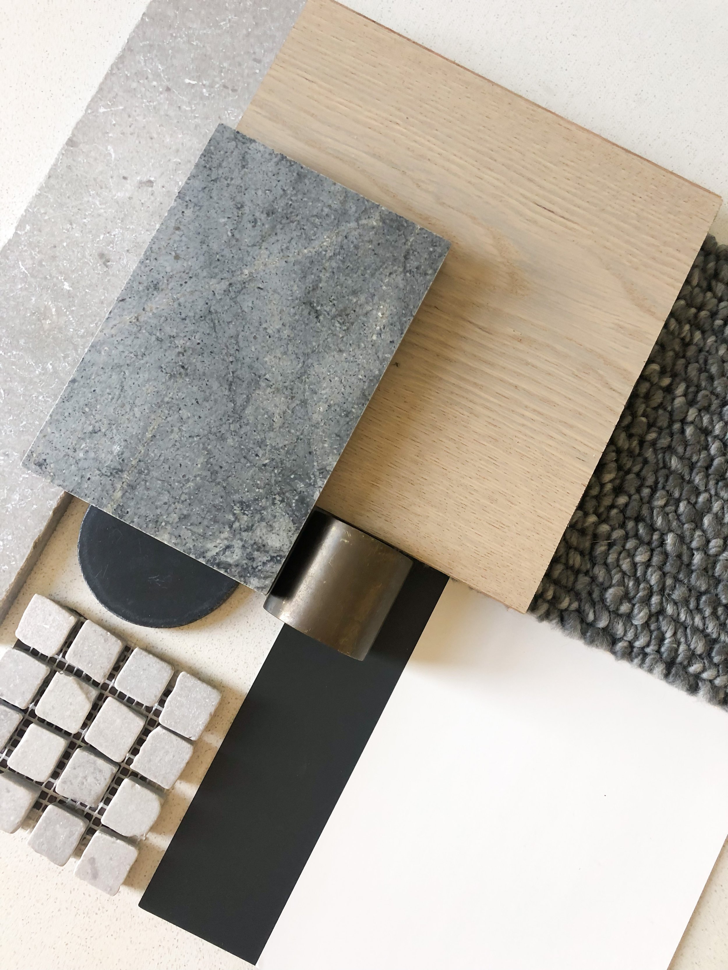 Sample materials for interior design, including tiles, a wooden panel, a plush gray rug, a stone slab, a black strip, and a metallic cylindrical piece.