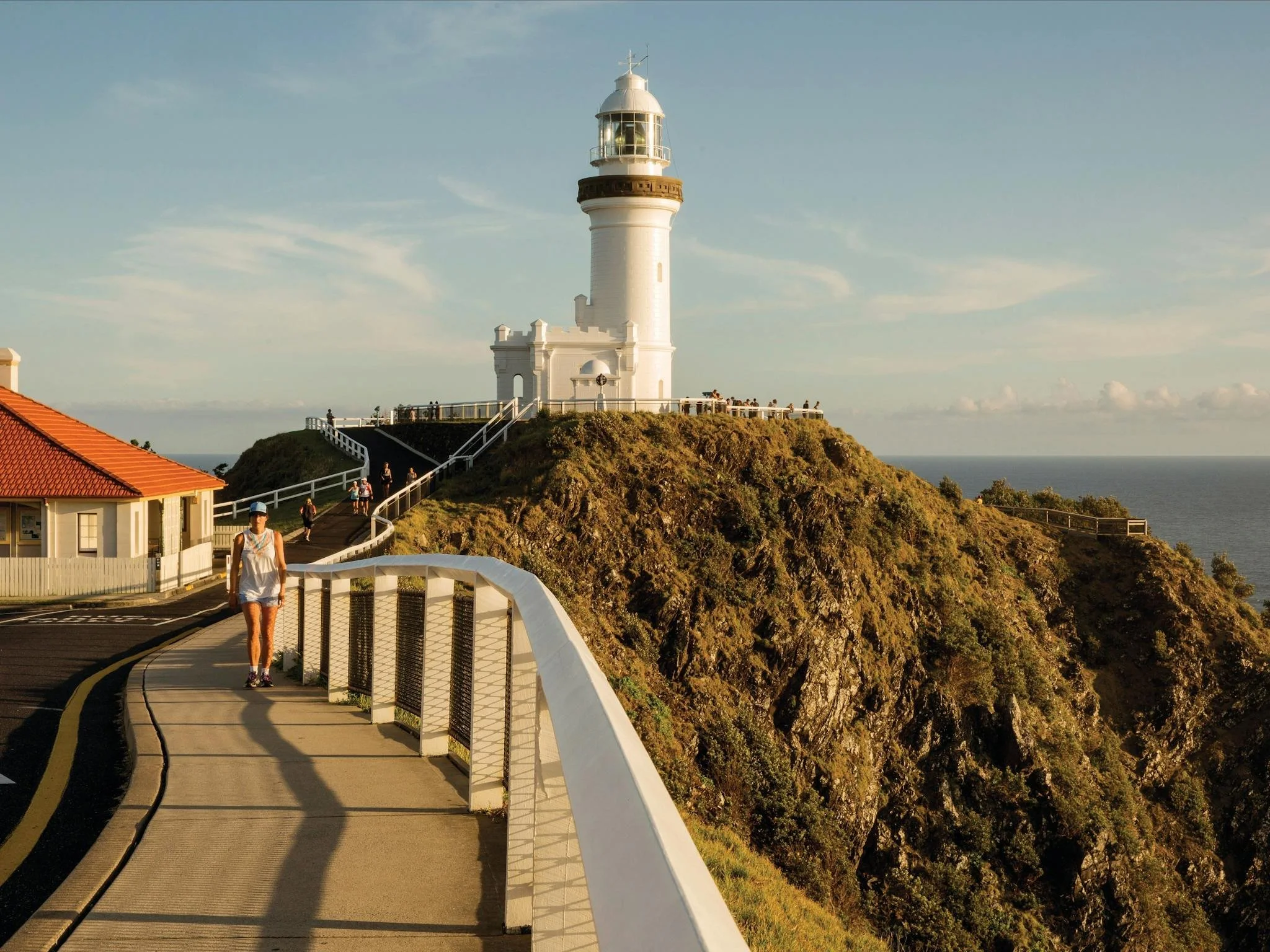 Pathway leading to a white lighthouse on a hill overlooking the ocean, with people walking nearby during sunset.