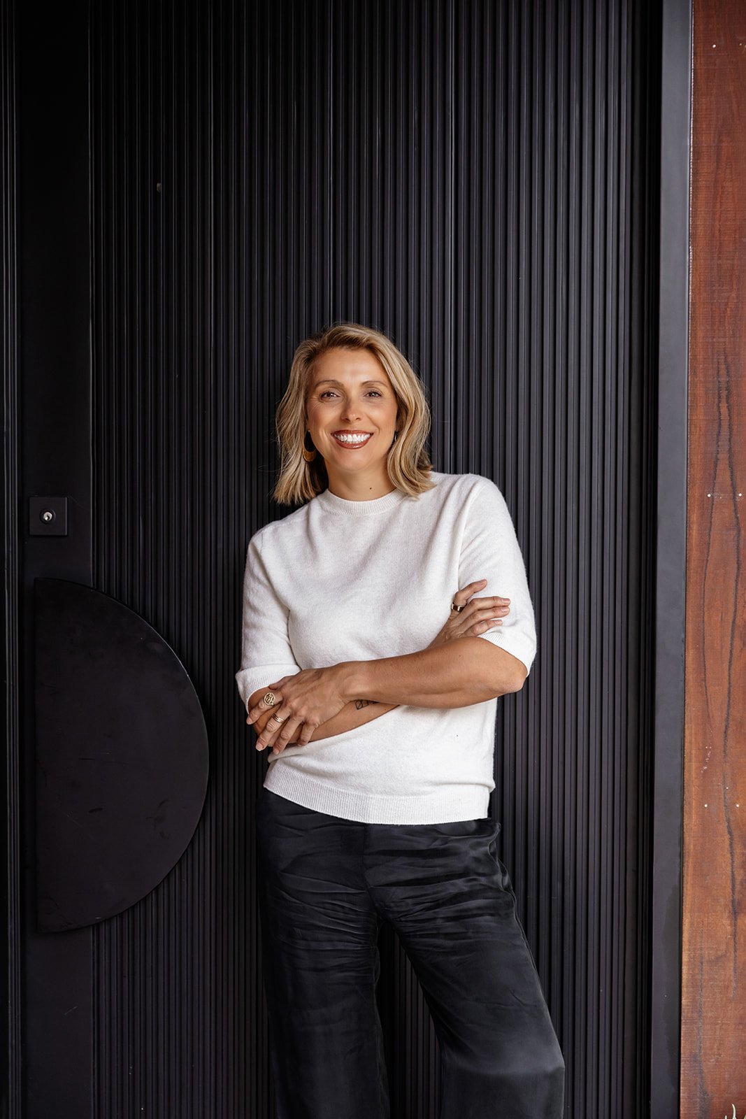 A woman with blonde, shoulder-length hair, smiling, wearing a white sweater and black pants, standing with arms crossed in front of a black textured wall.