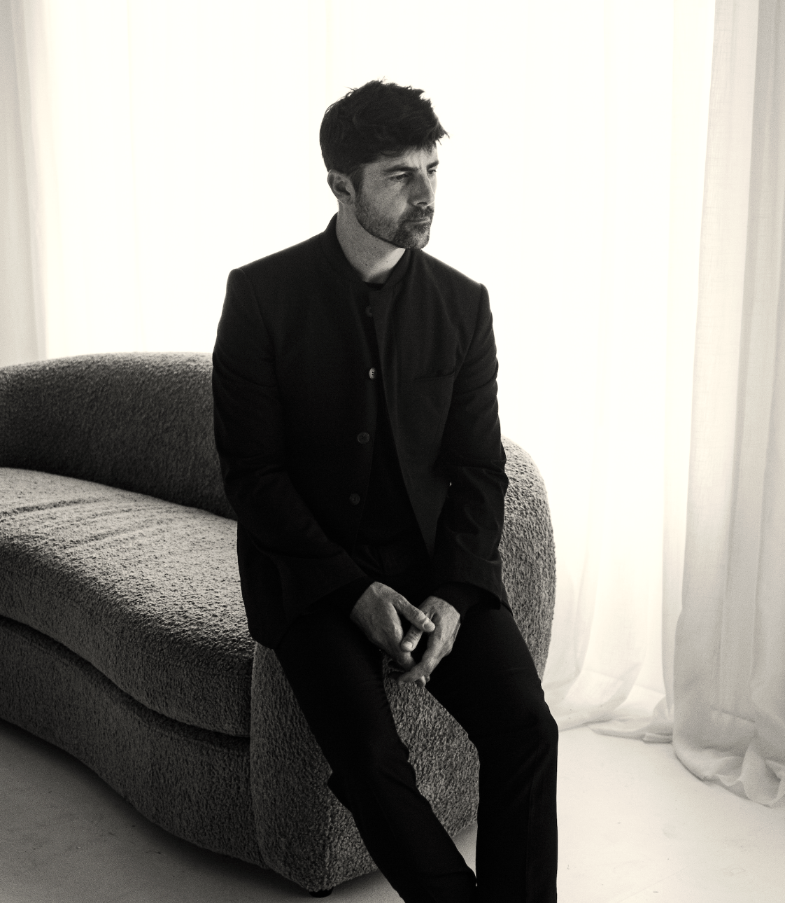 A man with dark hair and beard in a dark jacket sitting on a textured sofa in front of a bright, backlit window with curtains, looking pensively to the side.