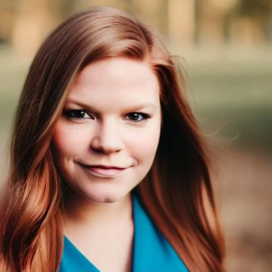 Close-up of a woman with long red hair, smiling softly, wearing a teal top, outdoors with blurred background.