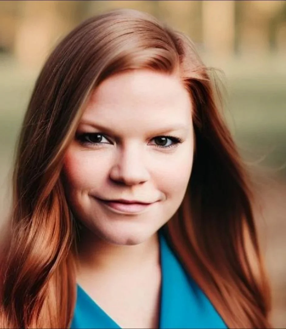 A young woman with long red hair smiling outdoors with a blurred natural background.