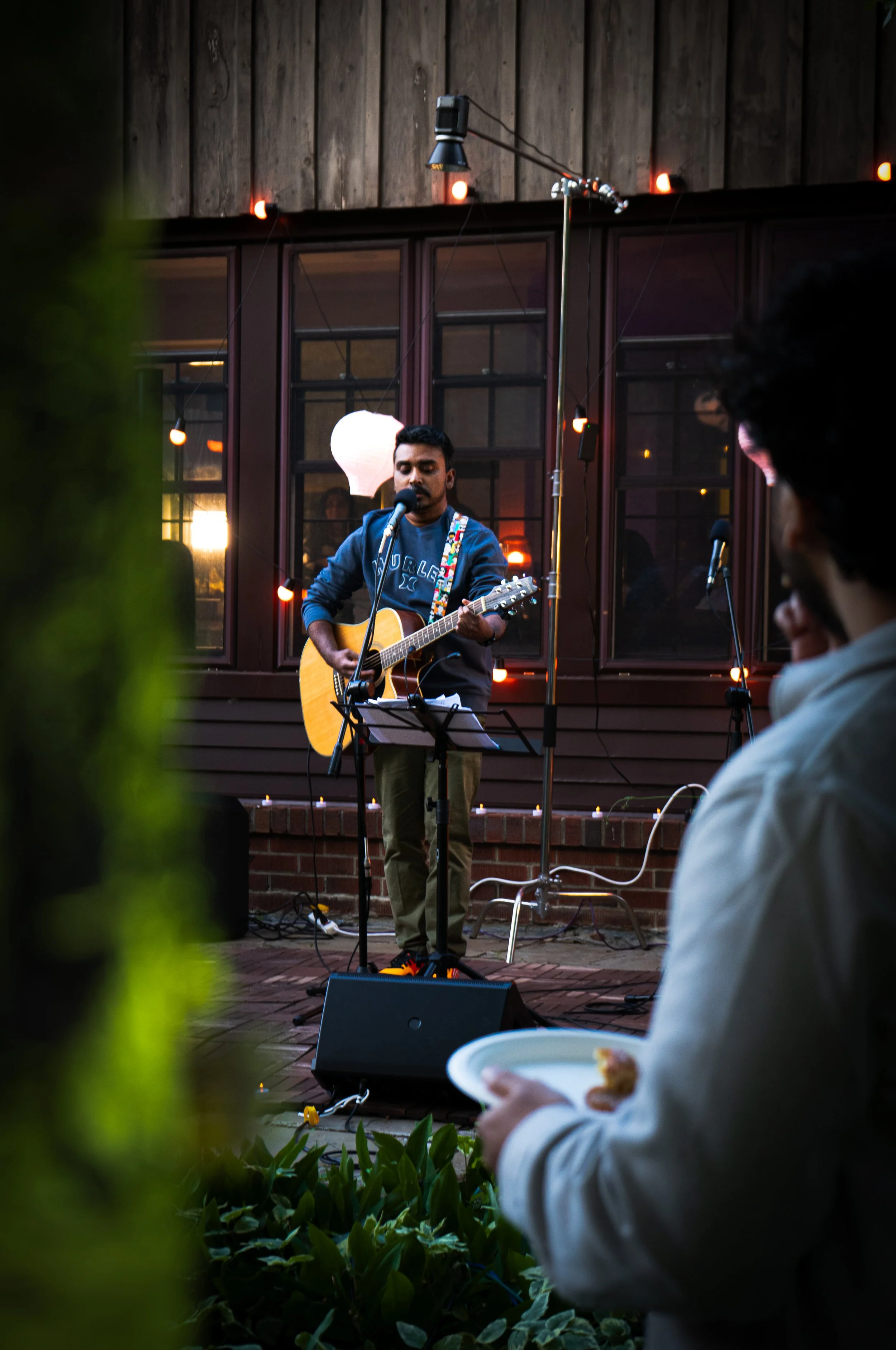 A man playing acoustic guitar and singing into a microphone outdoors during a live music performance, with an audience member in the foreground holding a plate of food, set against a wooden building with string lights.