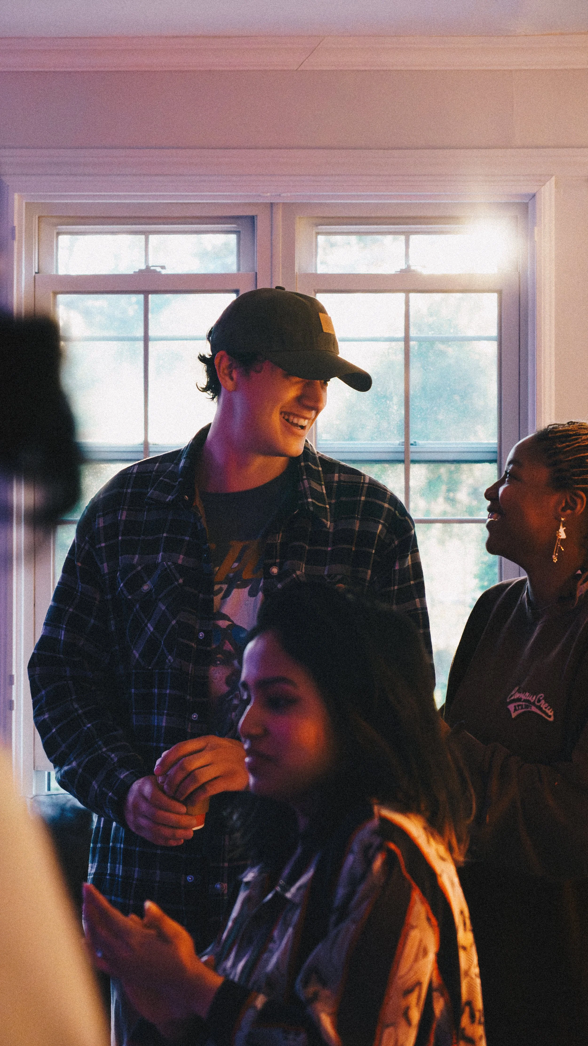 A group of people indoors near a window, with two women and a man smiling and interacting. The man is wearing a black baseball cap and a plaid shirt. One woman is looking at her phone, and the other woman is smiling, wearing earrings and a dark jacke