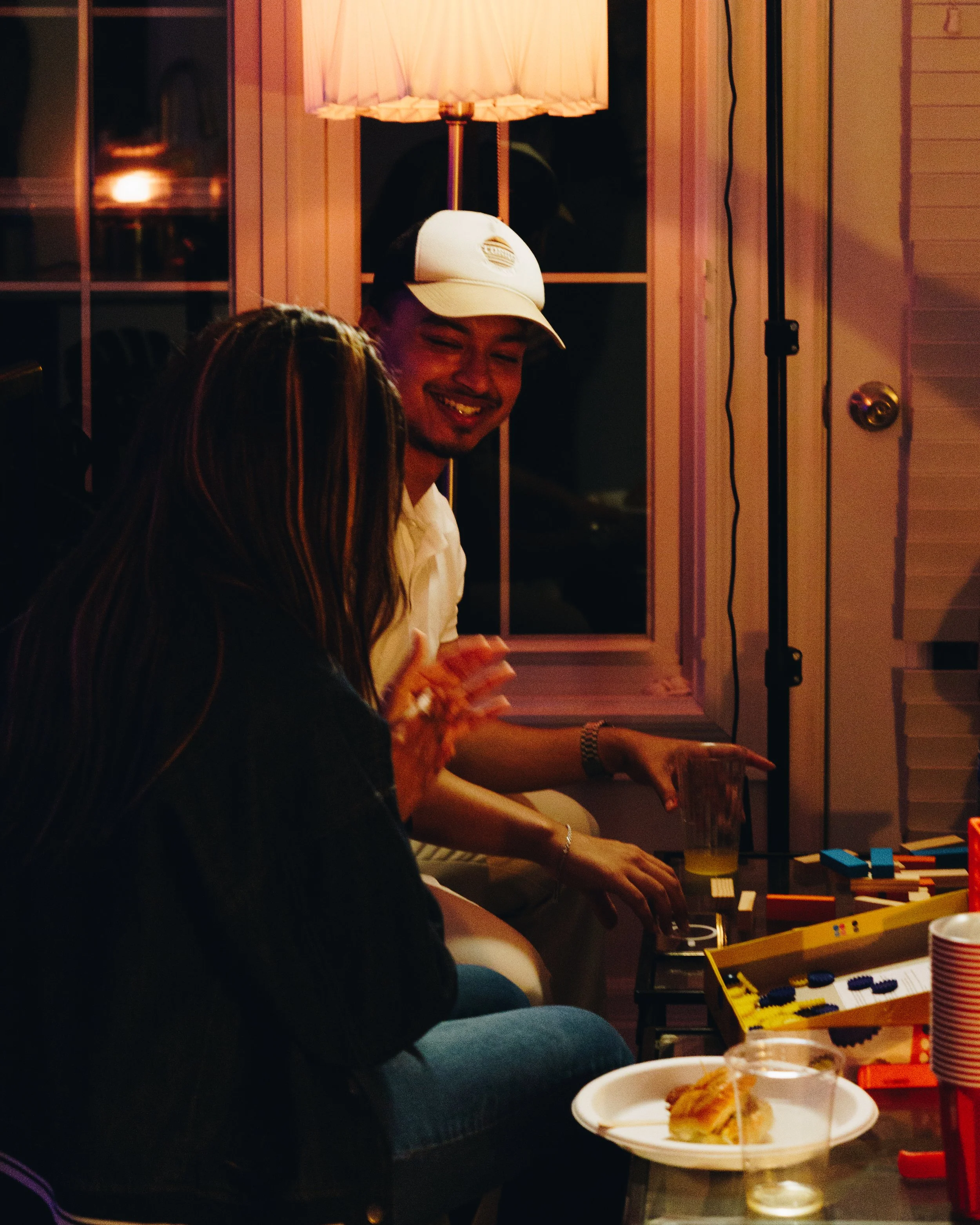 Two people sitting on a couch playing a board game, with snacks and drinks on the table, in a cozy, warmly lit room at night.