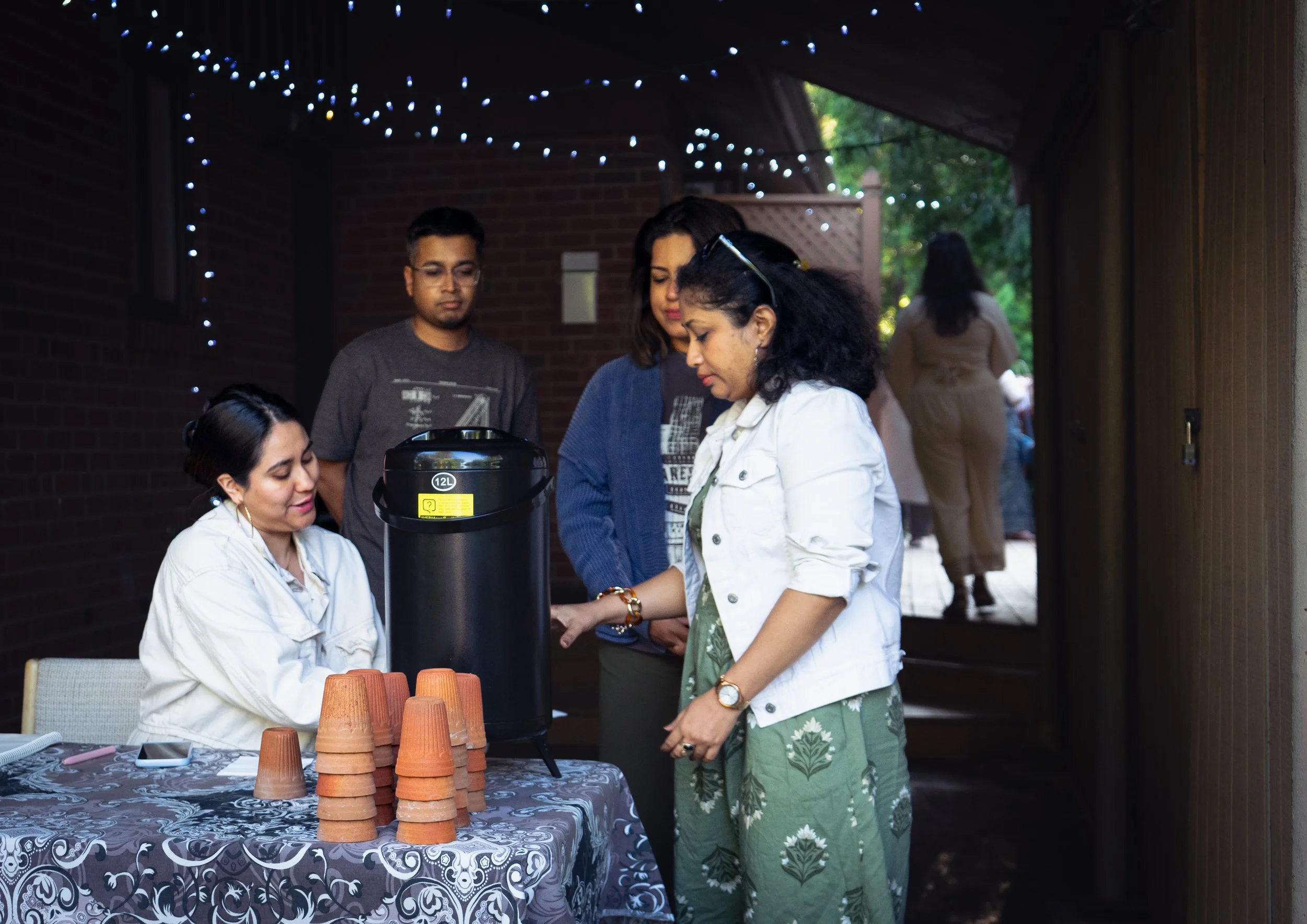 People gathered around a table with stacked terracotta pots, enjoying hot beverages from a large black thermos in a cozy outdoor setting decorated with string lights.
