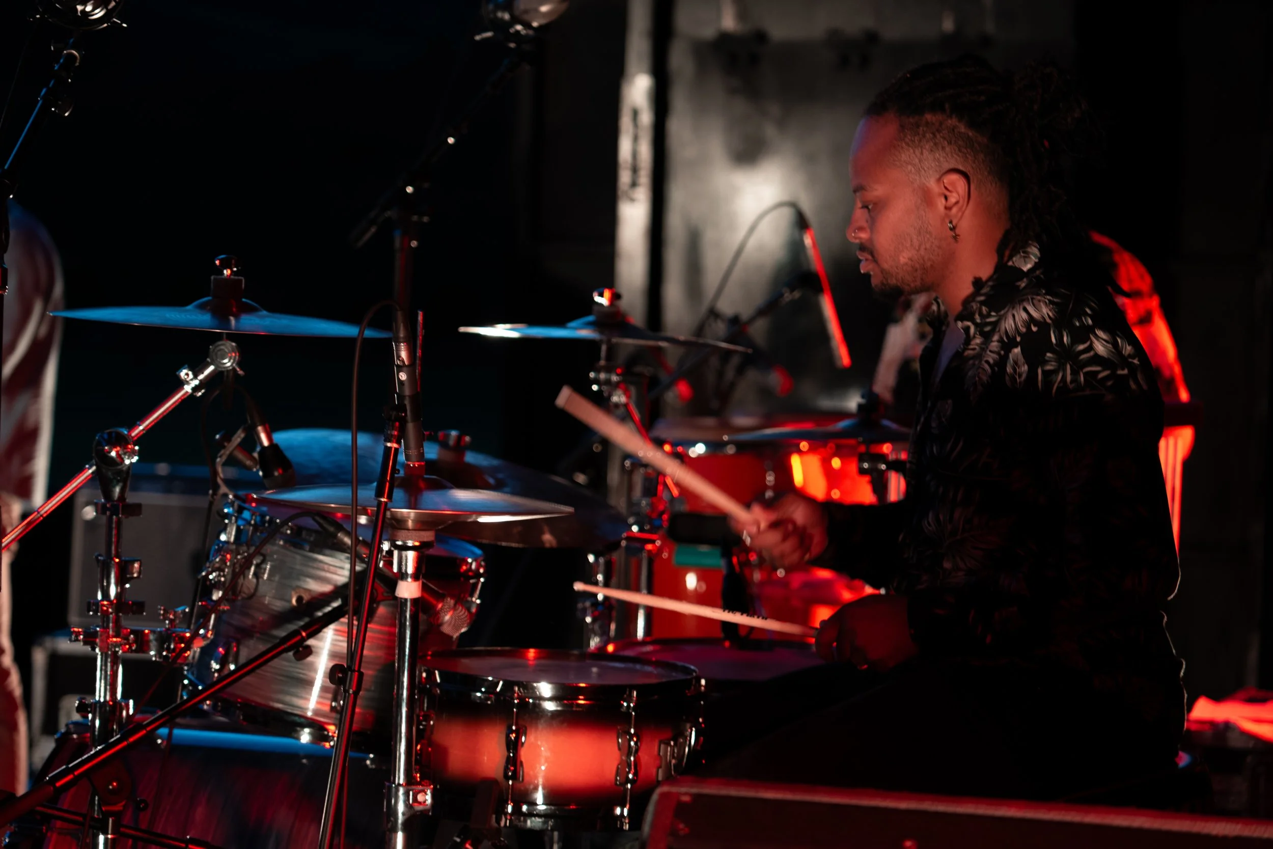 A male drummer with dreadlocks playing drums on a dimly lit stage, illuminated by red and blue stage lights. Jazz In the Park Church Road.