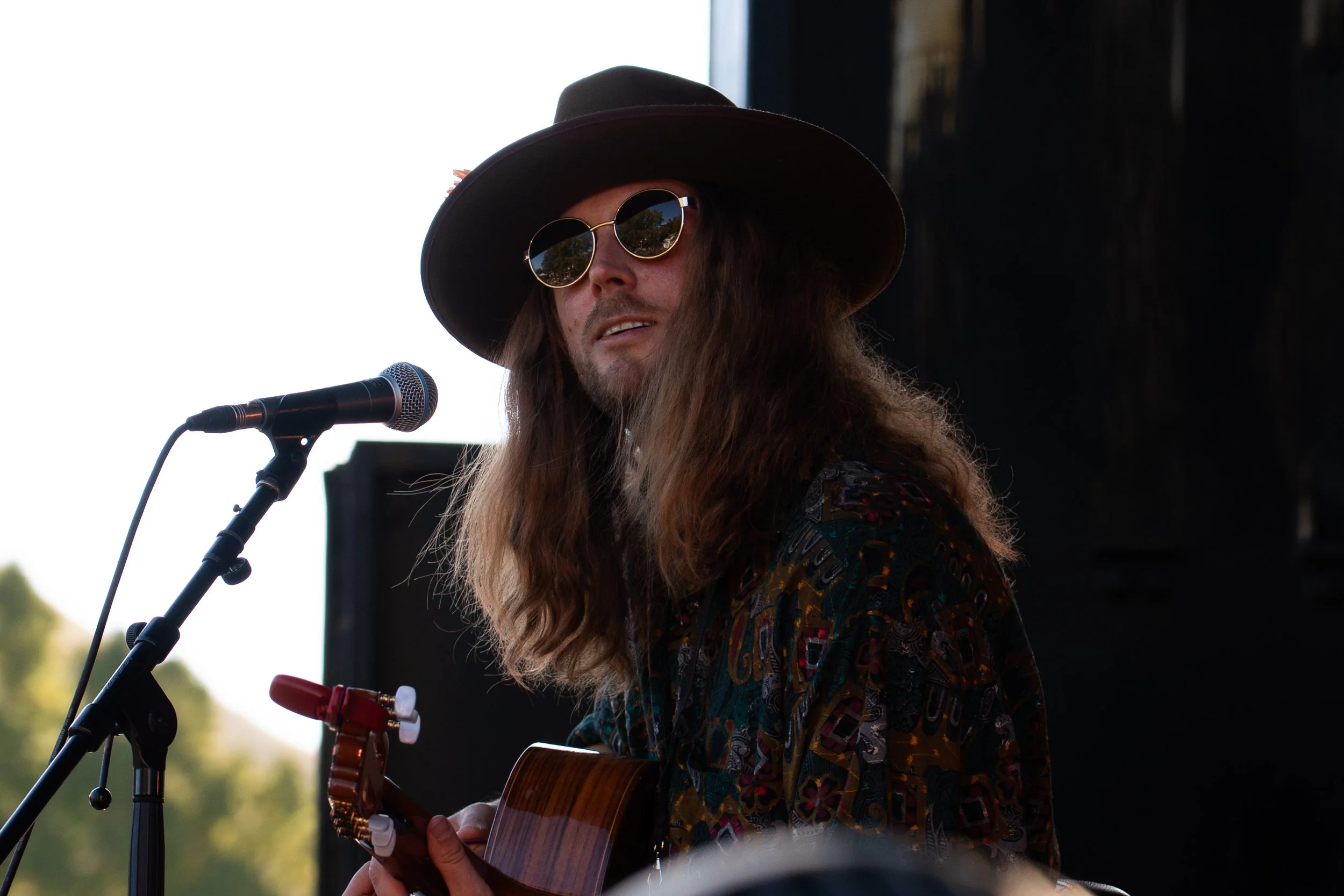 A man with long hair, wearing sunglasses, a wide-brimmed hat, and a patterned shirt, is playing an acoustic guitar and singing into a microphone at an outdoor event. Whiskey Green Horse - Jazz in the park, church road winery.