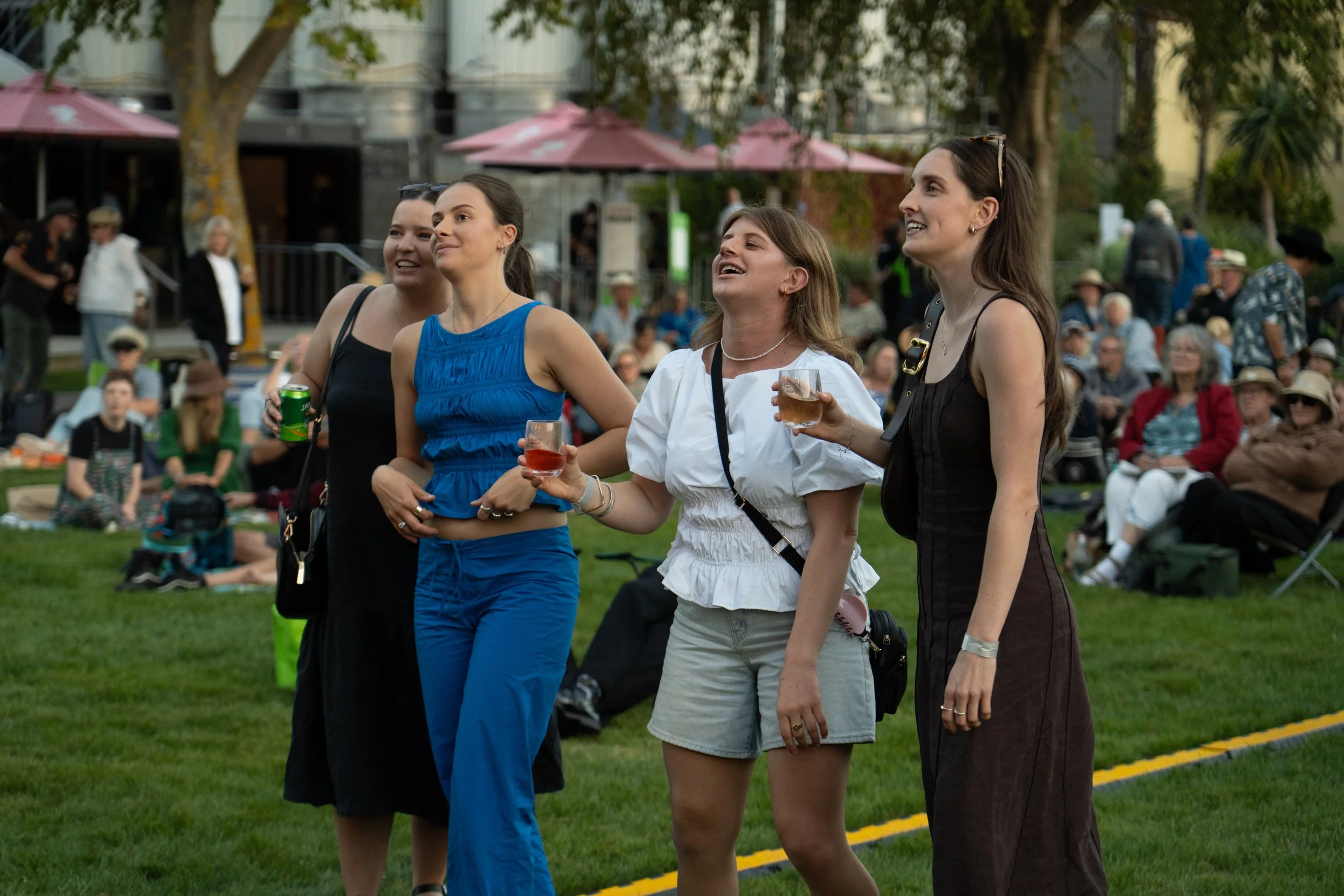 Four women stand together outdoors at a picnic, holding drinks and smiling, with a crowd seated on the grass behind them. Jazz in the park - church road winery.