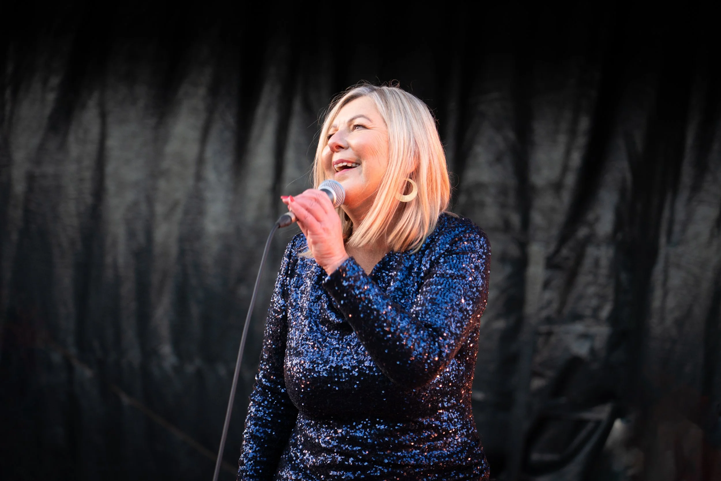 A woman singing into a microphone on stage, wearing a sparkly dark blue dress with a black background. Lady Larisa band - jazz in the park church road.