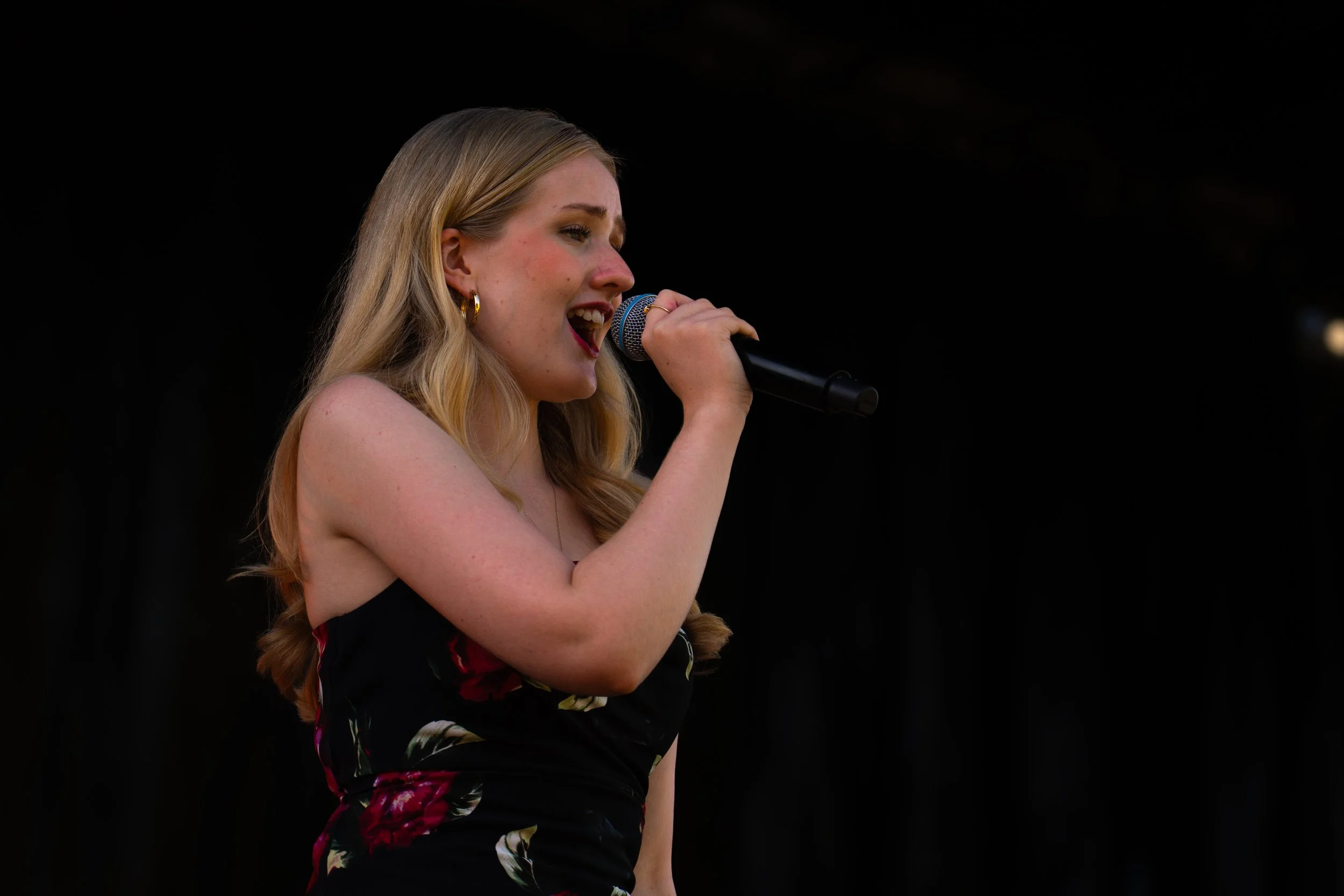 Young woman singing into a microphone on stage, wearing a black floral dress, with blonde hair and gold hoop earrings. Hawkes Bay Jazz Club Big Band. Jazz In the park church road winery.
