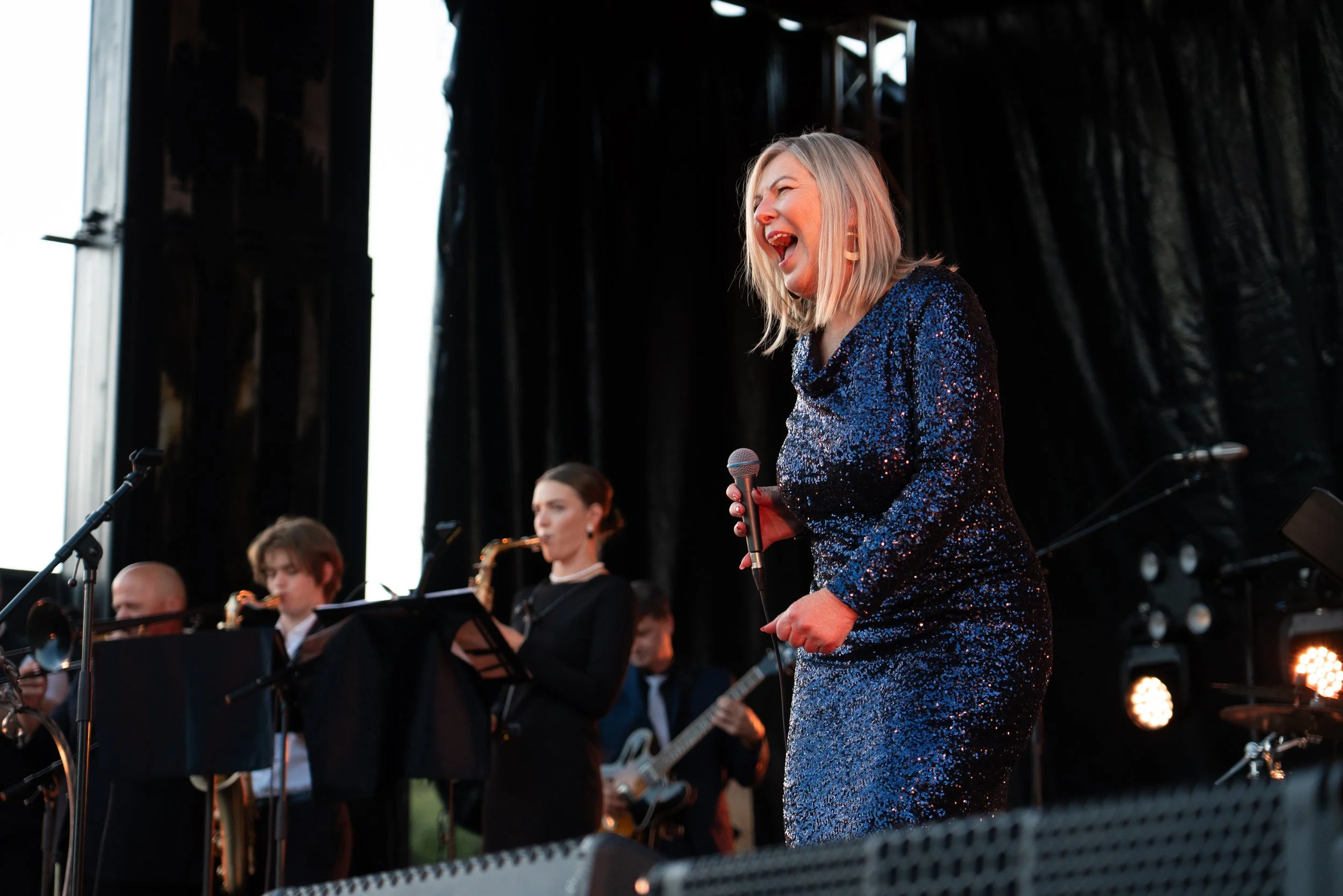 A woman in a sparkly blue dress passionately singing into a microphone on stage during a live jazz performance, accompanied by a band of saxophonists, guitarists, and a bassist. Lady Larisa band - jazz in the park church road.