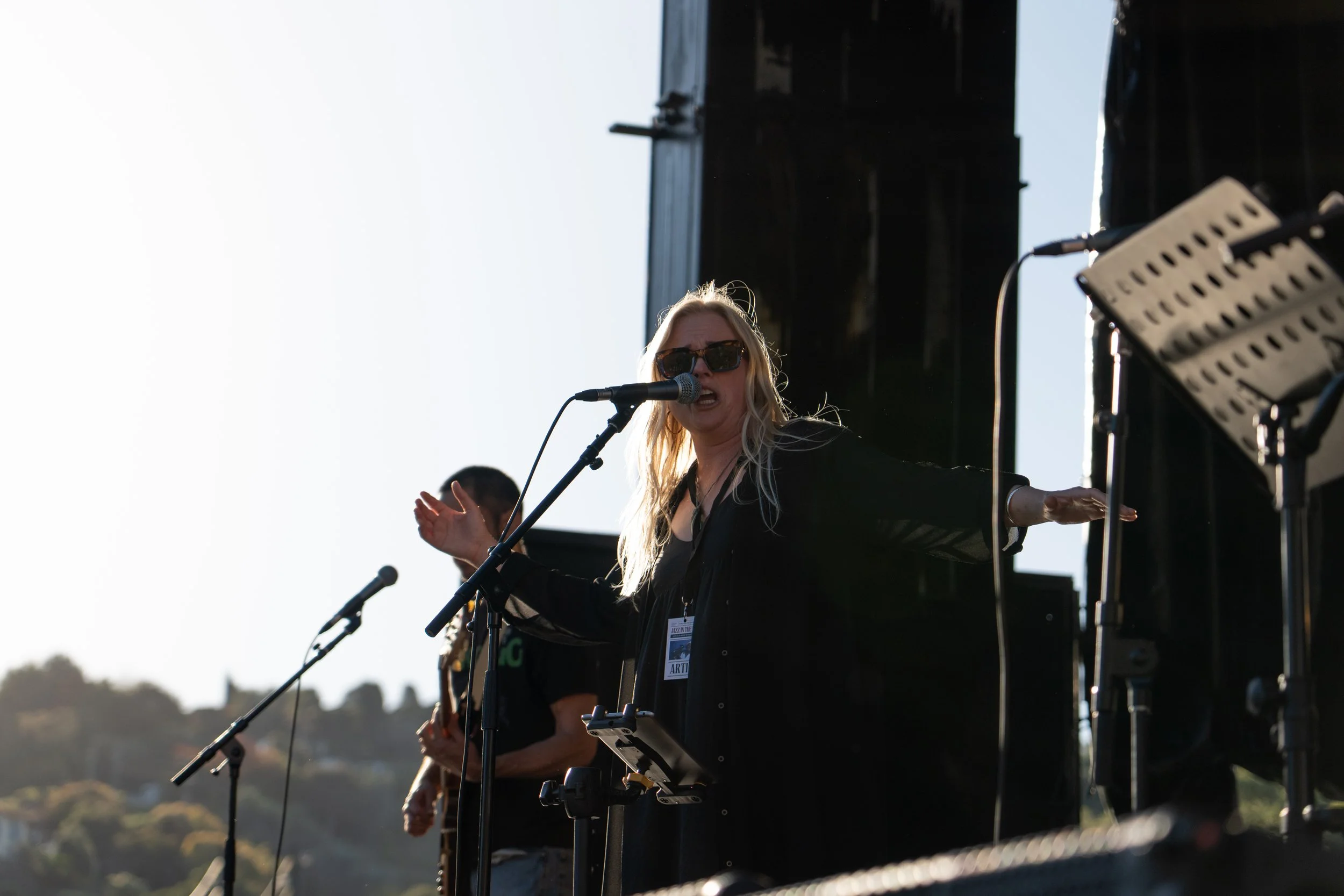 A female singer with blonde hair and sunglasses performs on a stage with microphones and musical equipment during an outdoor concert. Deep Fried Funk - jazz in the park church road winery.