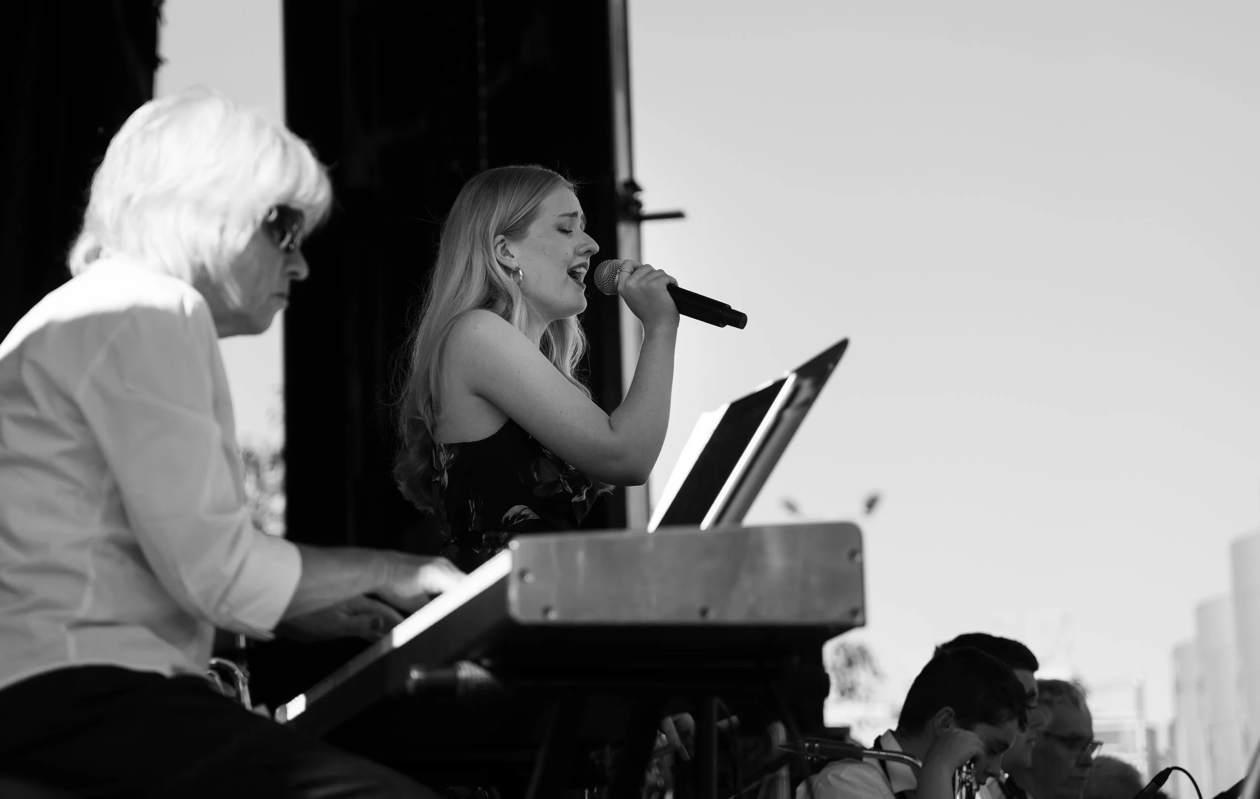 A woman singing into a microphone accompanied by a person playing a keyboard on stage, with other musicians visible in the background, during a live outdoor performance. Hawkes Bay Jazz Club Big Band. Jazz In the park church road winery.
