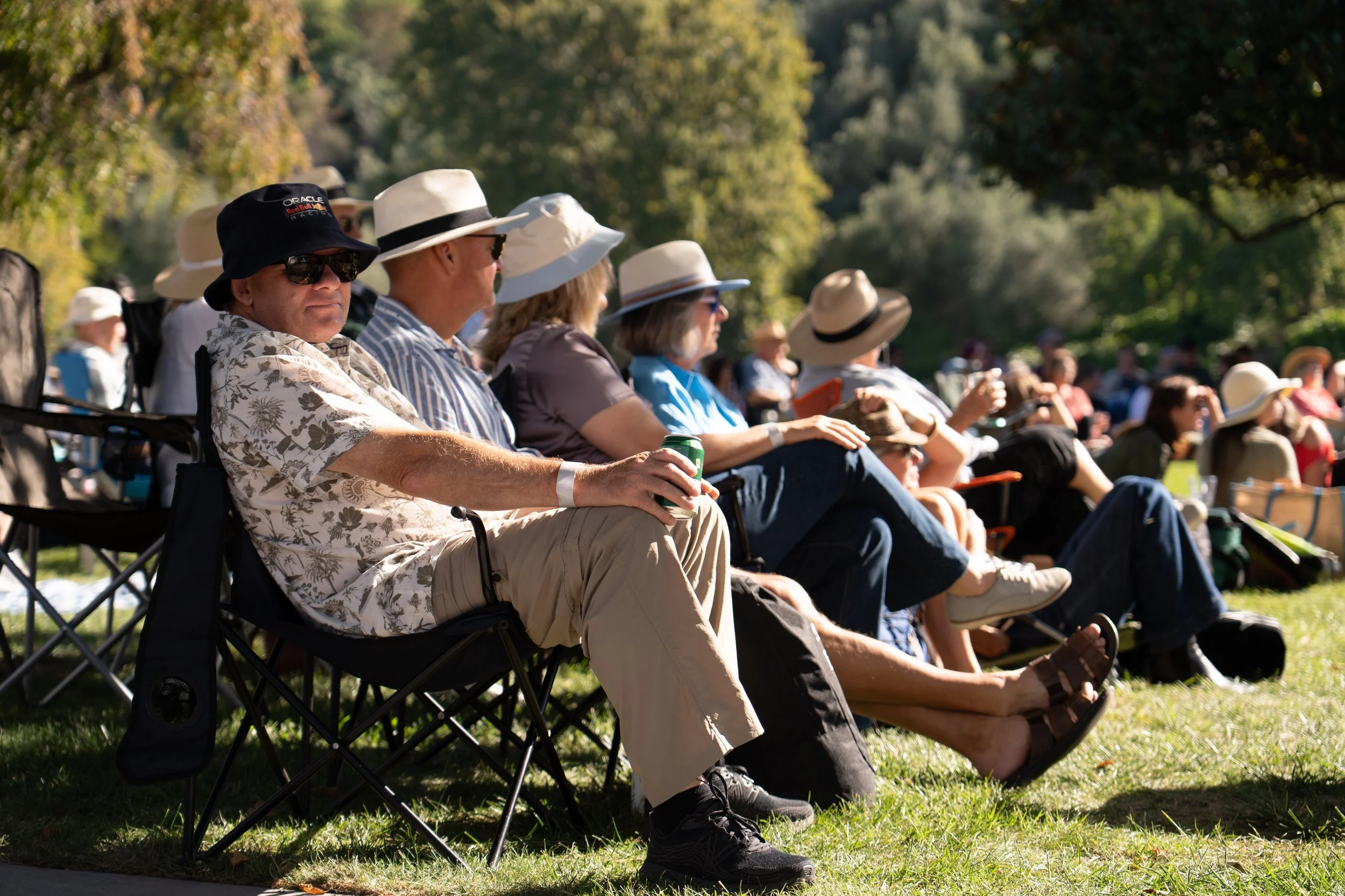 Group of people seated outdoors on folding chairs in a park, wearing hats and sunglasses on a sunny day. Jazz In the park church road winery.