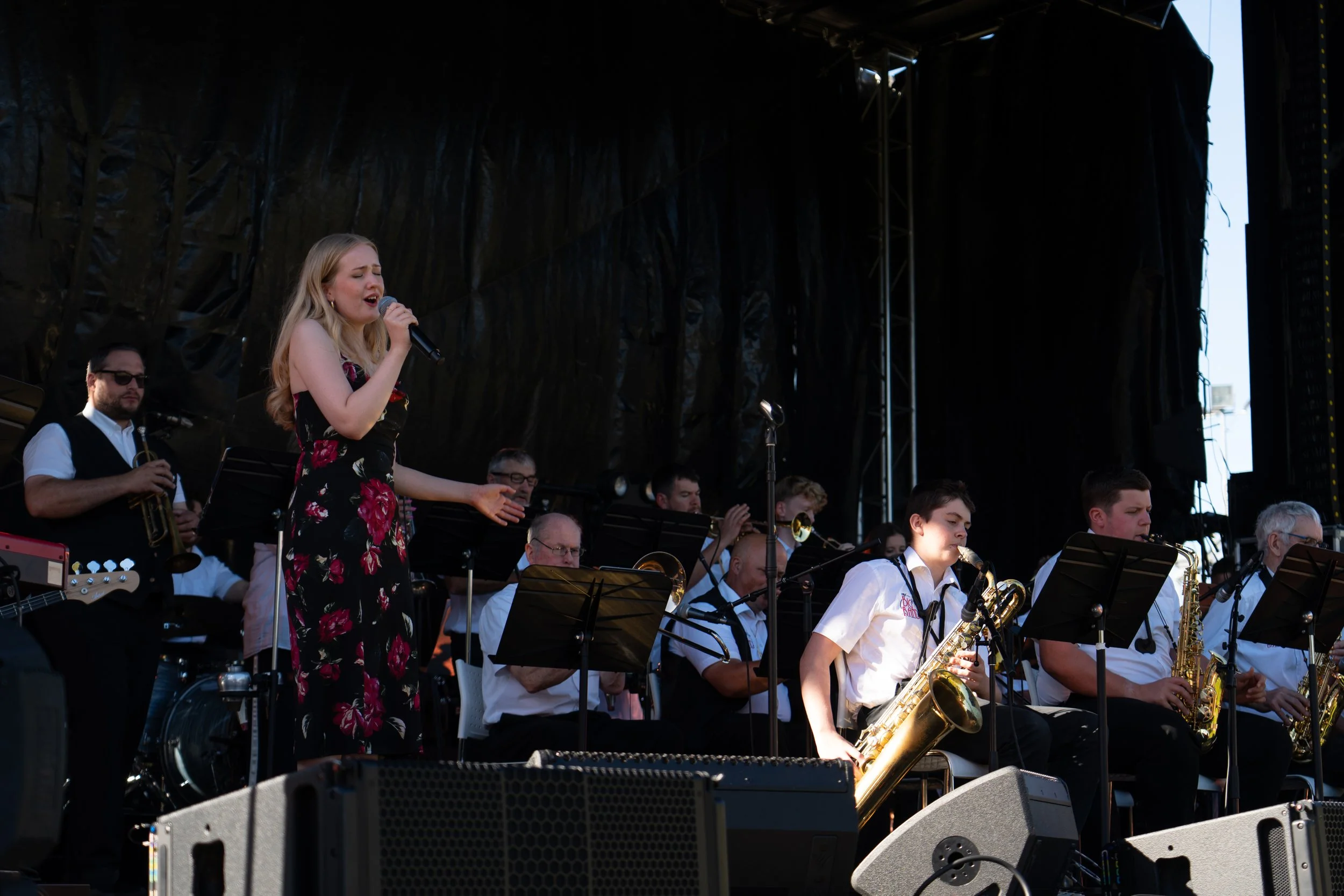 A woman singing into a microphone on stage with a band. Several young men playing saxophones and band members are visible behind her, some wearing white shirts and black vests. Hawkes Bay Jazz Club Big Band. Jazz In the park church road winery.