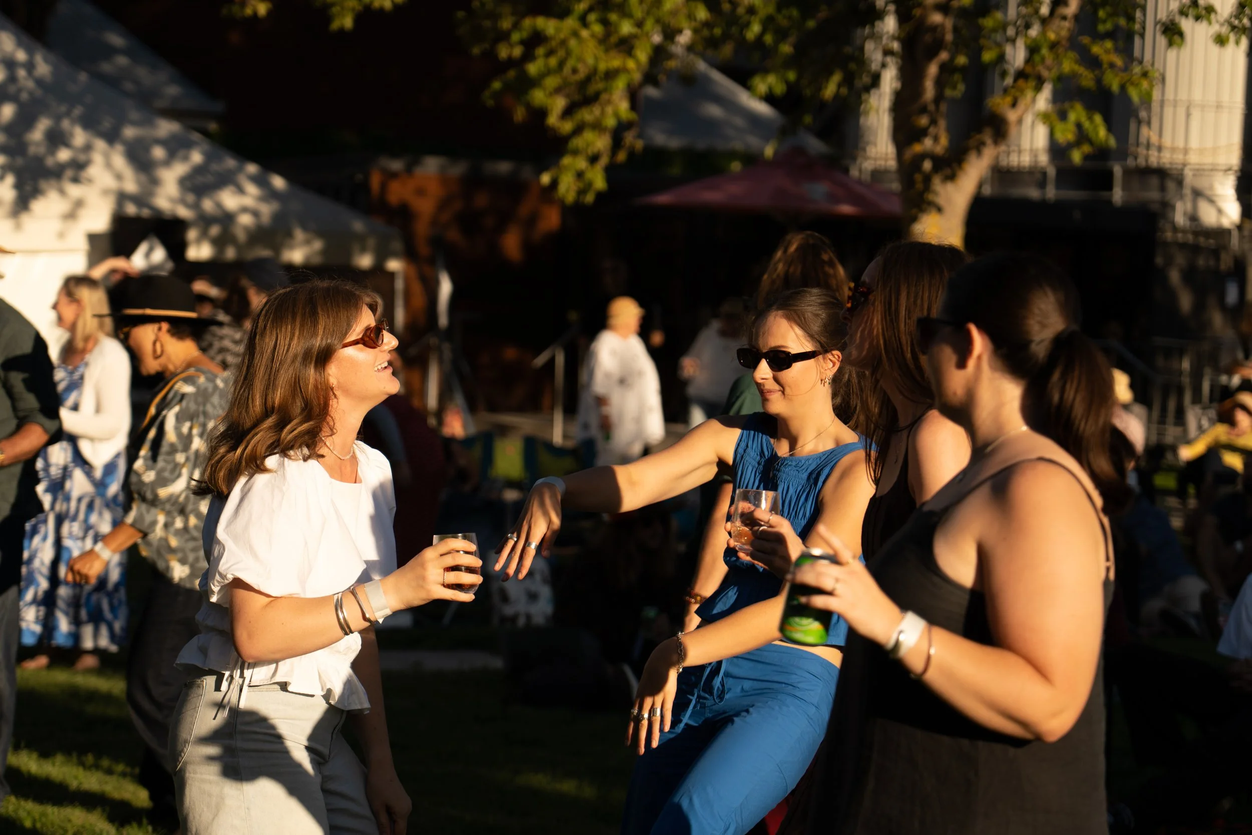 Group of women socializing outdoors in the evening, wearing sunglasses and holding drinks, at a festive gathering with tents and trees in the background. Jazz in the park church road winery.