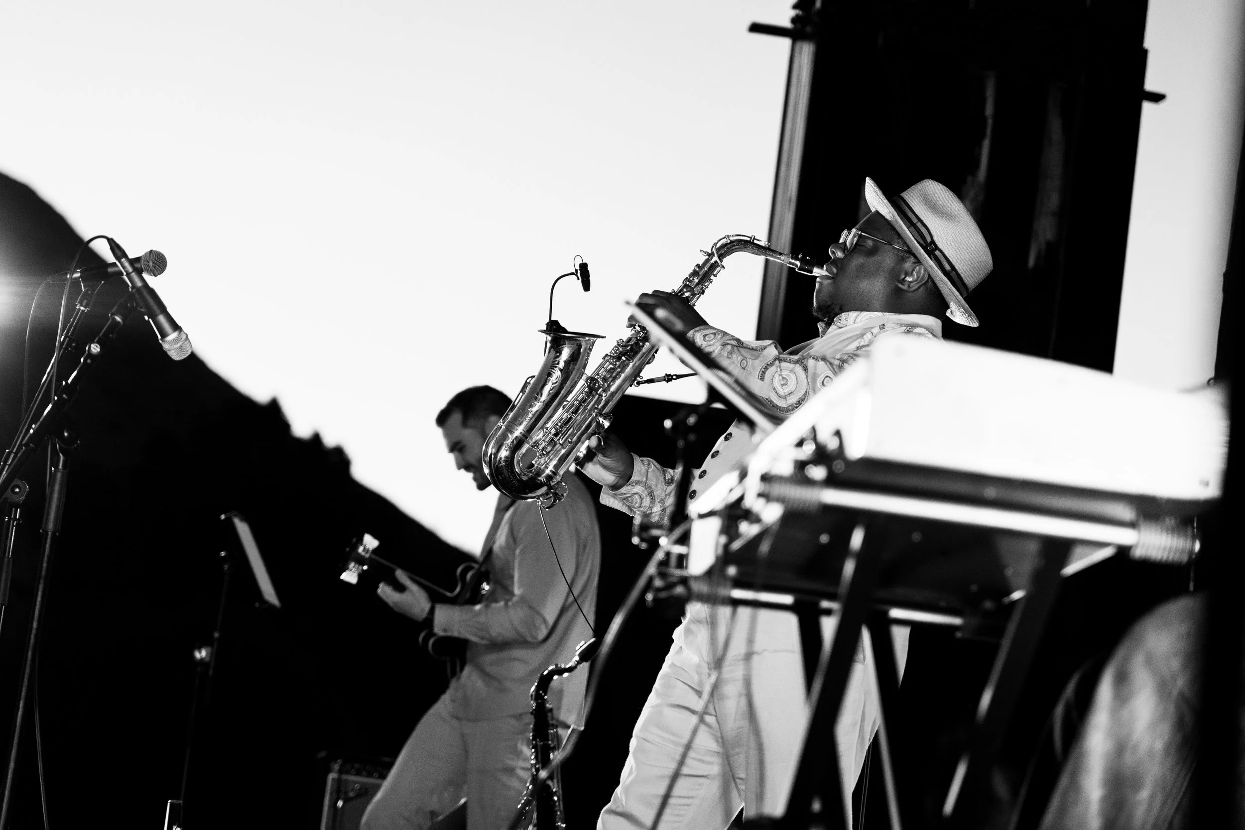 Two musicians performing on stage: one playing a keyboard and the other playing a saxophone, wearing a hat and glasses, in a black and white photograph. Thabani Gapara band - jazz in the park church road.