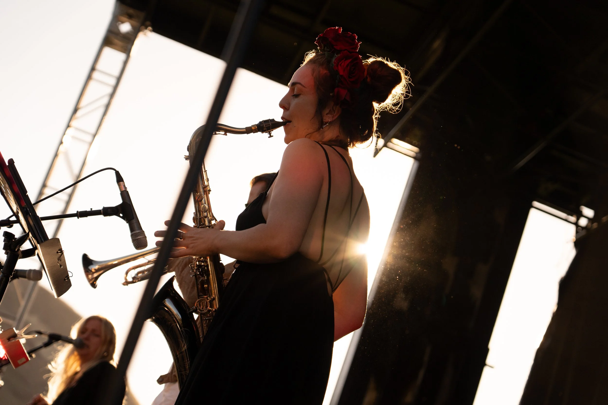 A woman with a floral headpiece playing a saxophone on stage at sunset. Deep Fried Funk - jazz in the park church road winery.