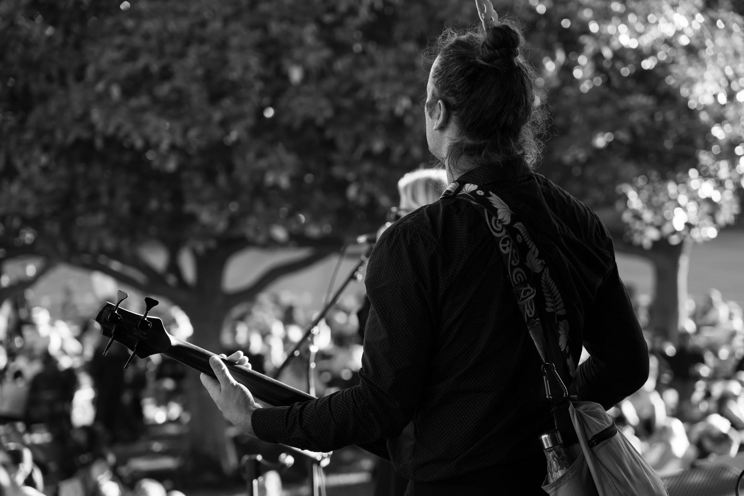 A person with a bun hairstyle holding a guitar outdoors during daytime, with large trees and a crowd in the background. Deep Fried Funk - jazz in the park church road winery.