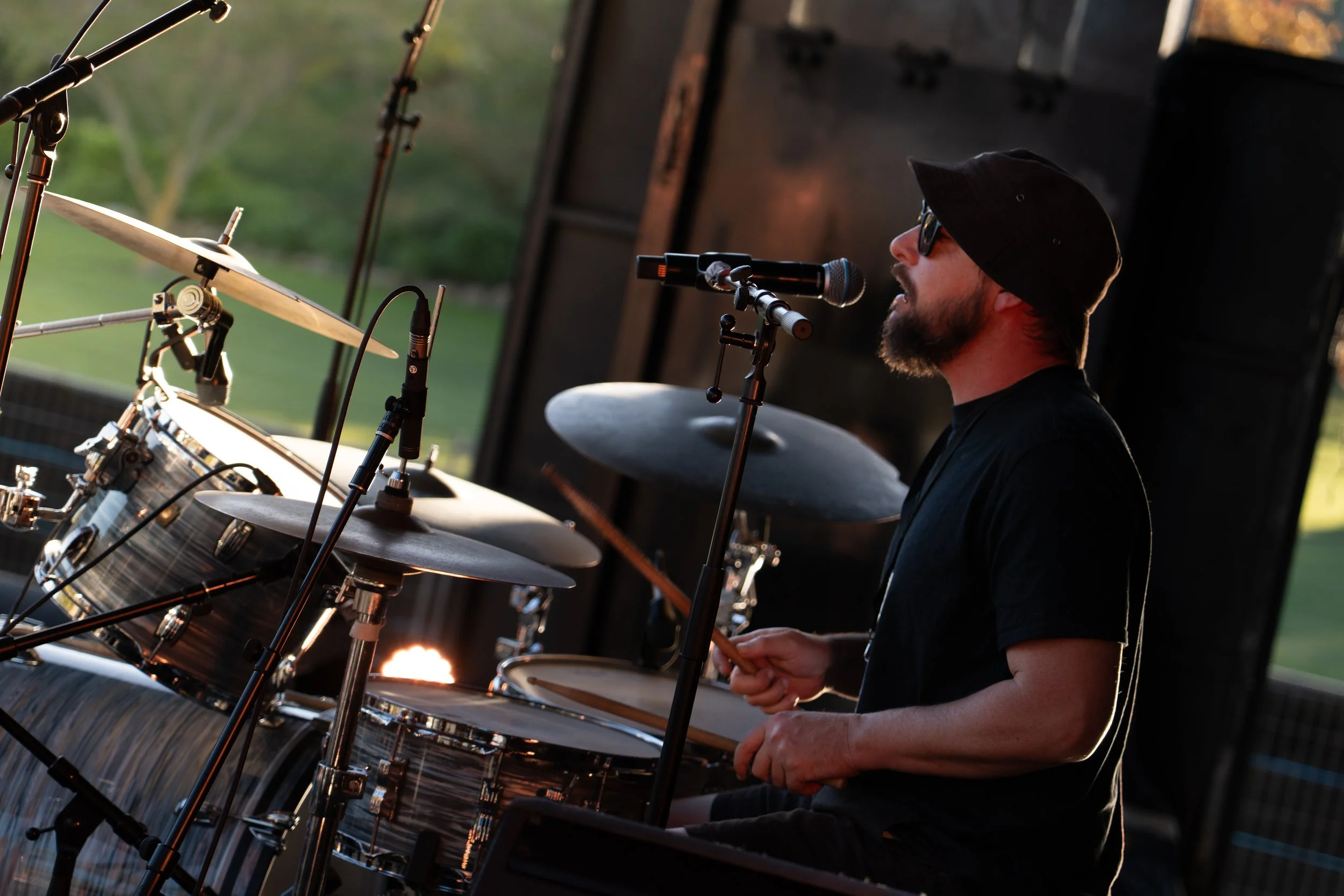 A musician wearing a black hat and sunglasses playing drums on stage with a microphone in front of him during an outdoor performance at sunset. Deep Fried Funk - jazz in the park church road winery.