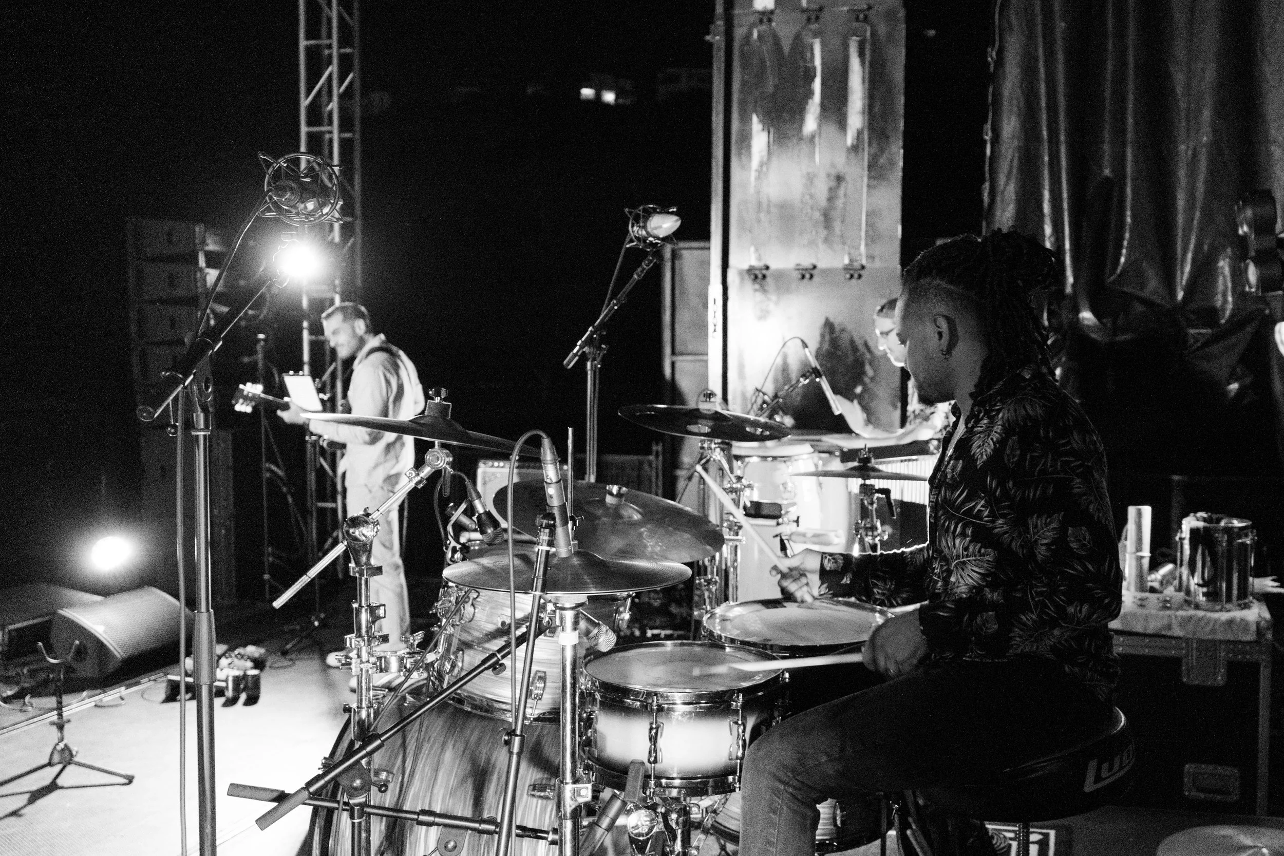 A black and white photo of thabani gapara band performing on stage at jazz in the park church road. A drummer with dreadlocks is playing a drum set, and a guitarist and another band member are visible in the background.