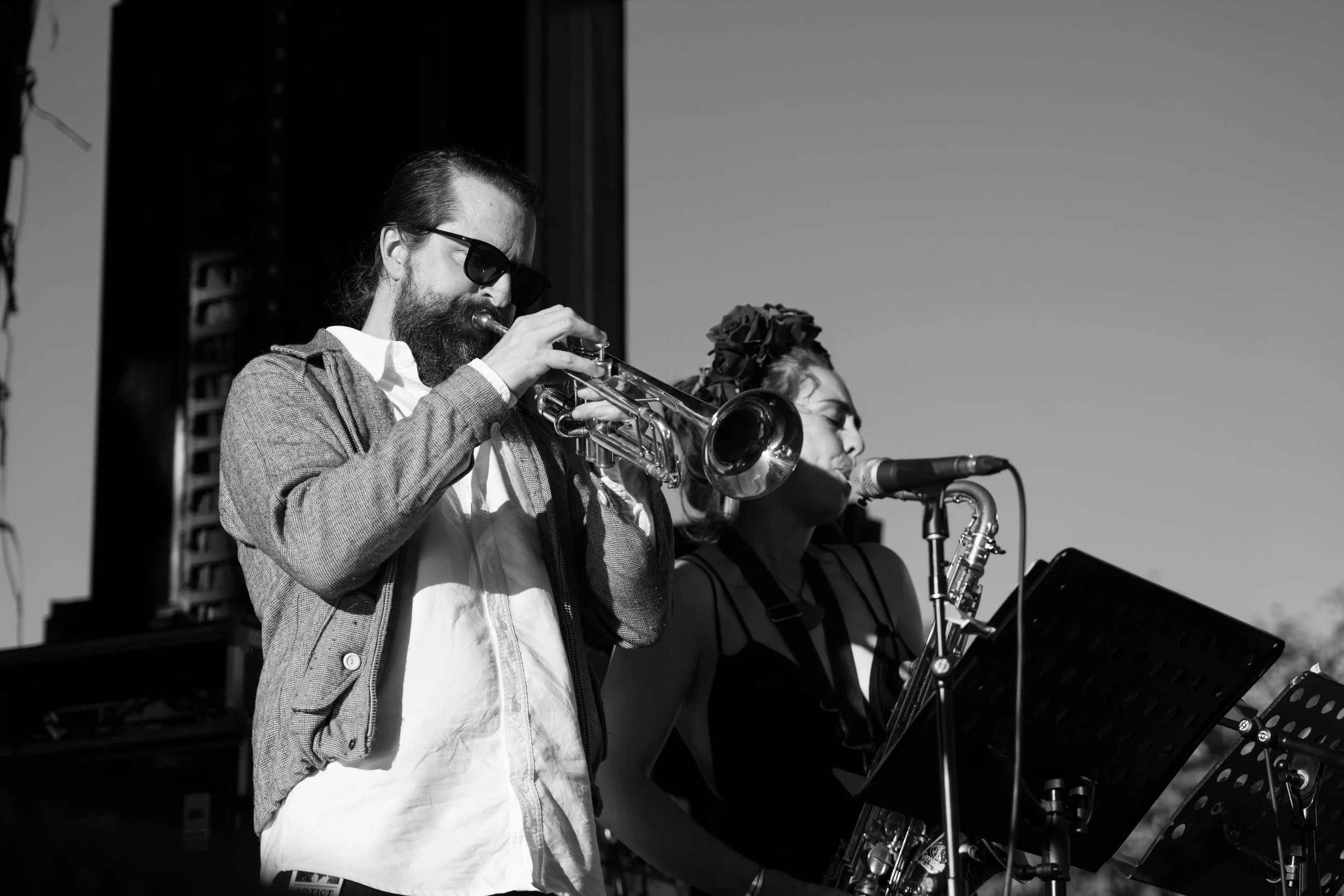 Black and white photo of a male musician with sunglasses playing trumpet and a female musician singing into a microphone during a performance. Deep Fried Funk - jazz in the park church road winery.