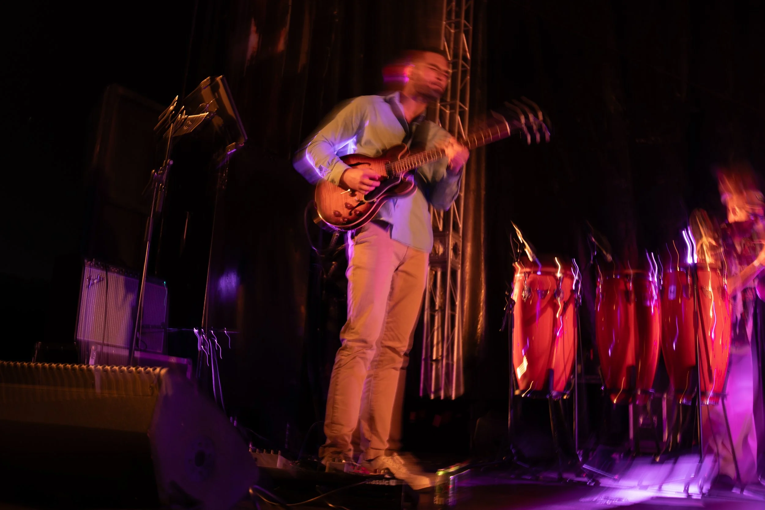 Musician playing an electric guitar on stage with red conga drums to the side, illuminated by colorful stage lights, with a blurred motion effect. Thabani Gapara band - jazz in the park church road.