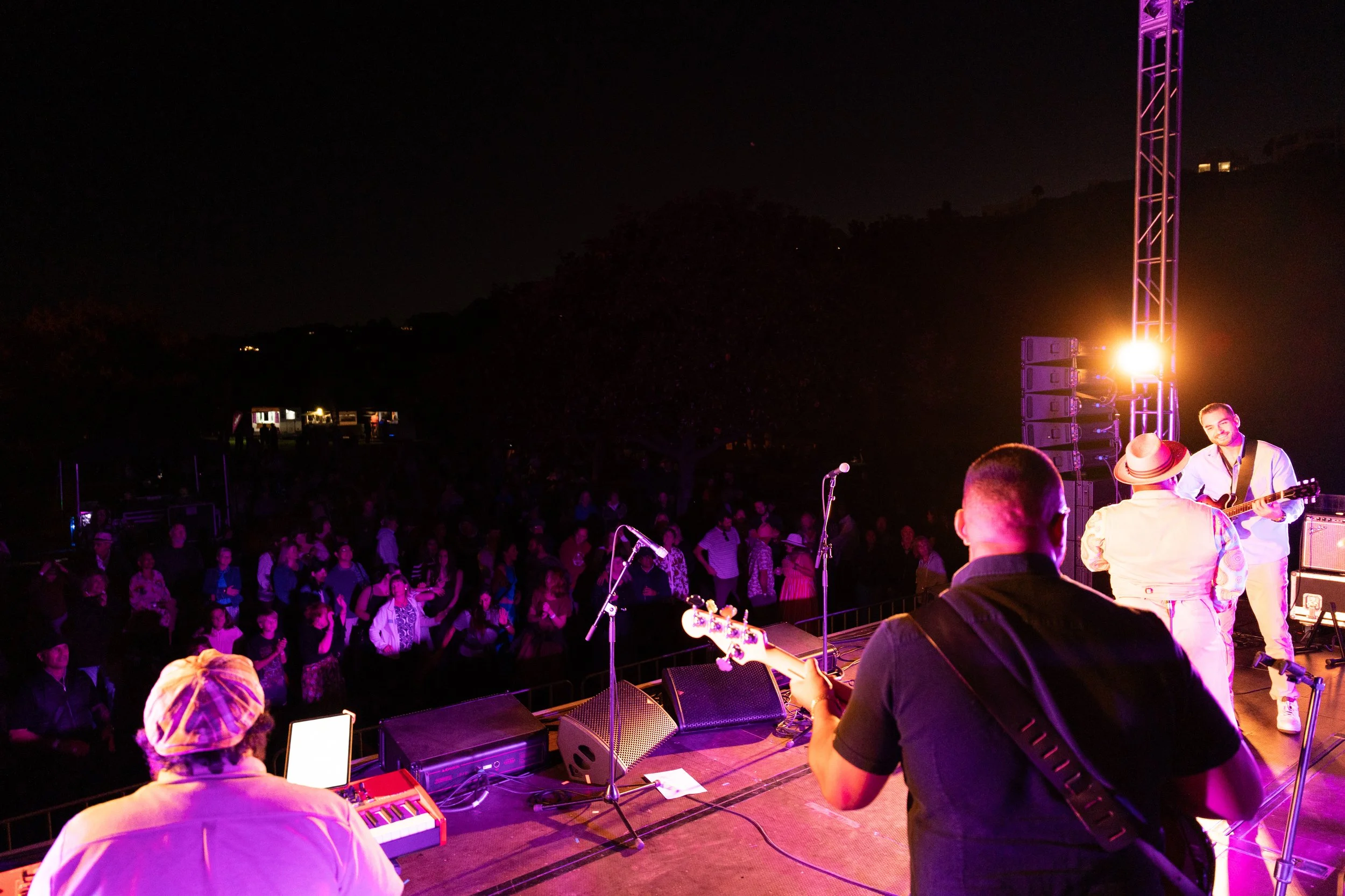 Musicians performing on stage at an outdoor night concert, with a crowd of people watching. Jazz in the park church road.