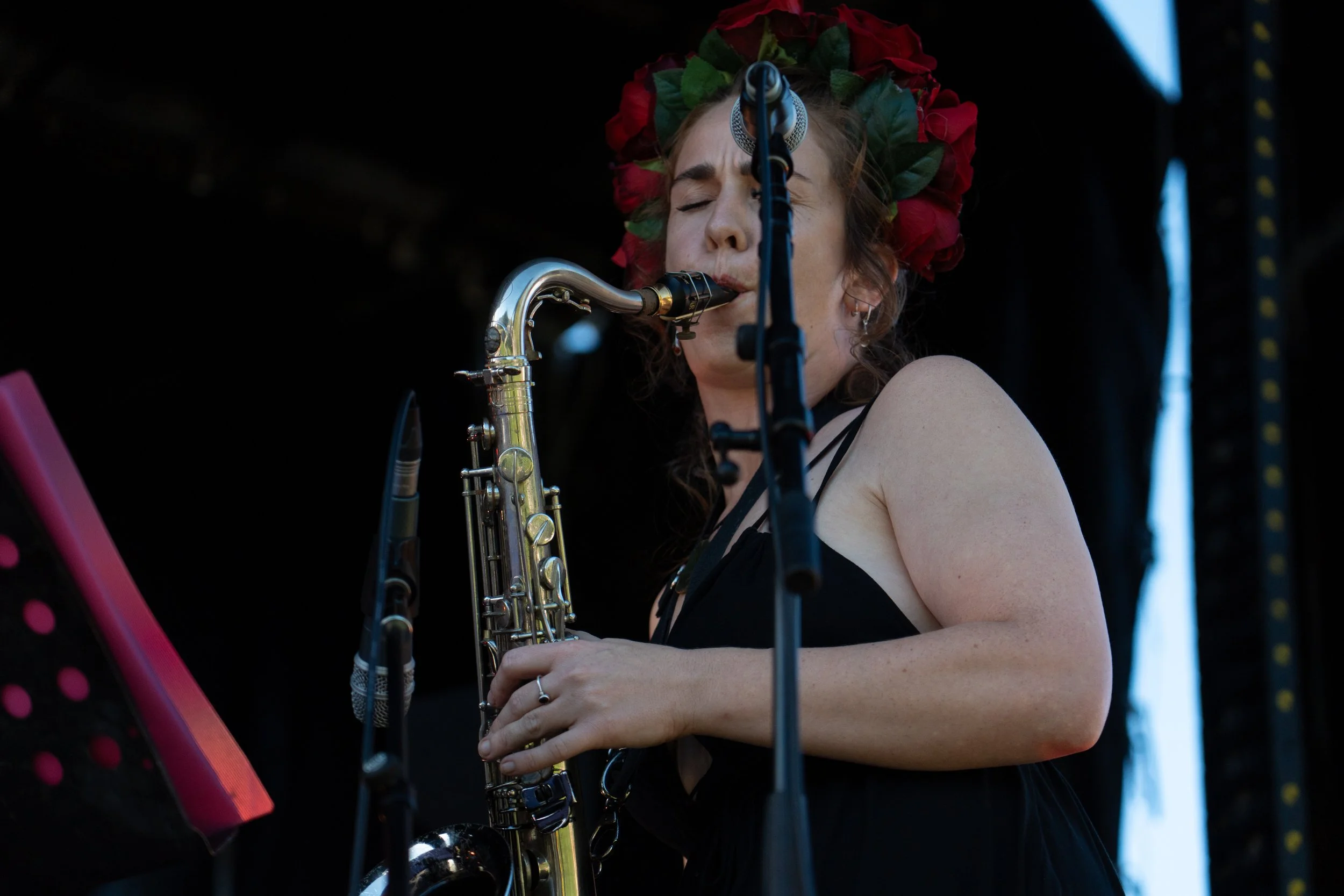 A woman with long, curly hair wearing a red flower crown and black dress playing a saxophone on stage, with a black background and presence of stage equipment. Whiskey Green Horse - Jazz in the park, church road winery.