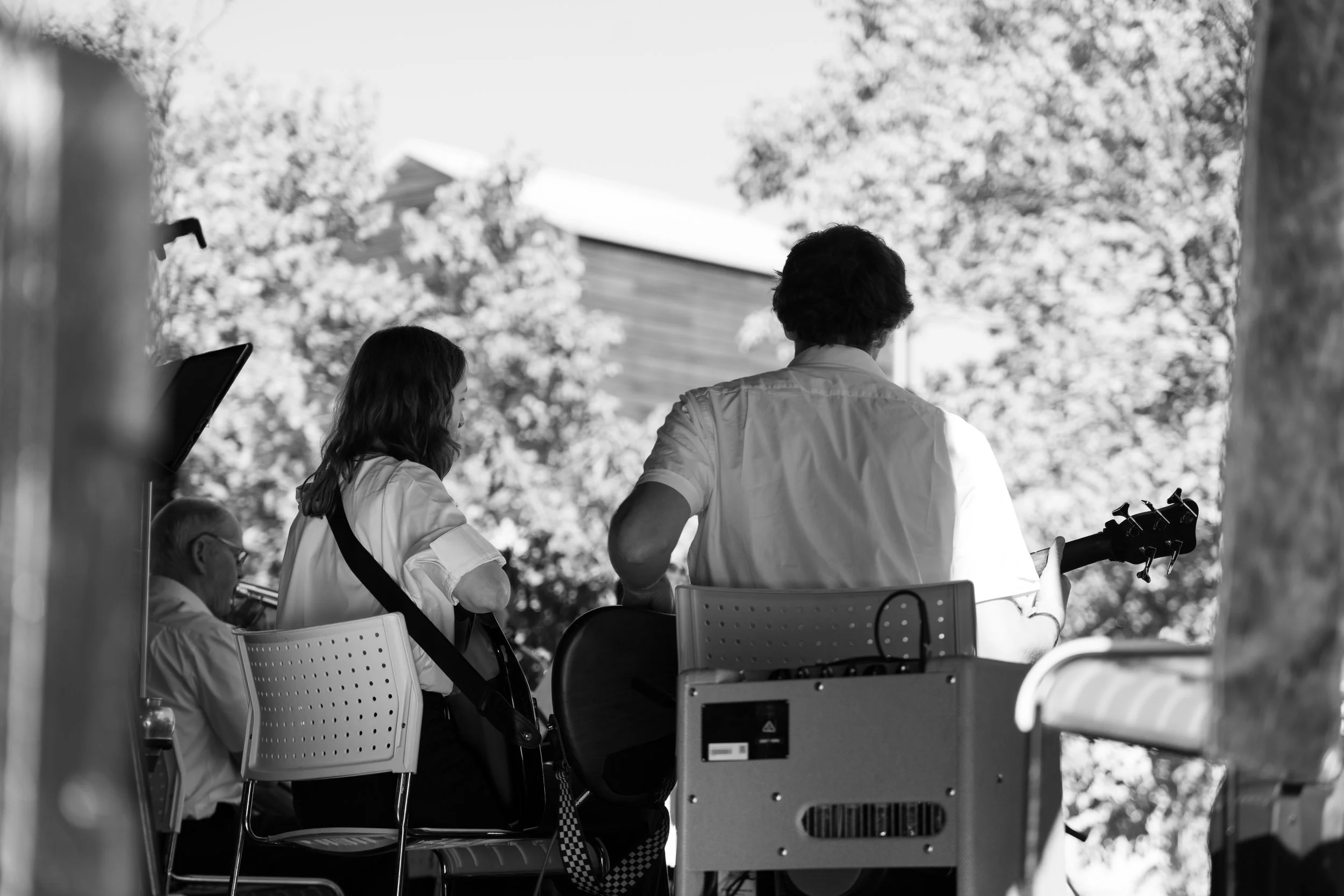 Black and white photo of a group of people performing music outdoors, with one person playing guitar, a woman with a violin, and an older man possibly playing a wind instrument. Hawkes Bay Jazz Club Big Band. Jazz In the park church road winery.