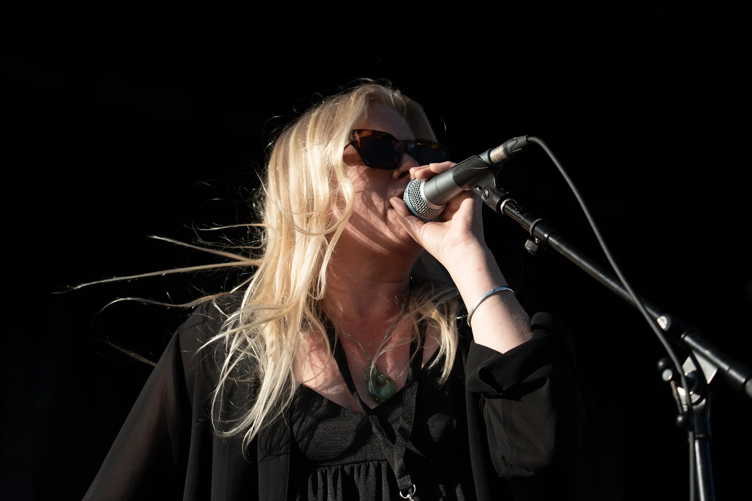 A woman with blonde hair, wearing sunglasses and a black top, singing into a microphone on a dark background. Deep Fried Funk - jazz in the park church road winery.