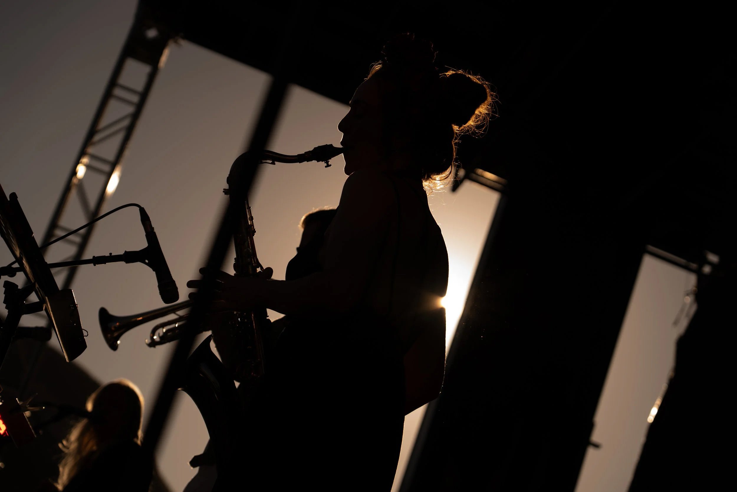 Silhouette of a woman playing the saxophone on stage with sunlight in the background. Deep Fried Funk - jazz in the park church road winery.
