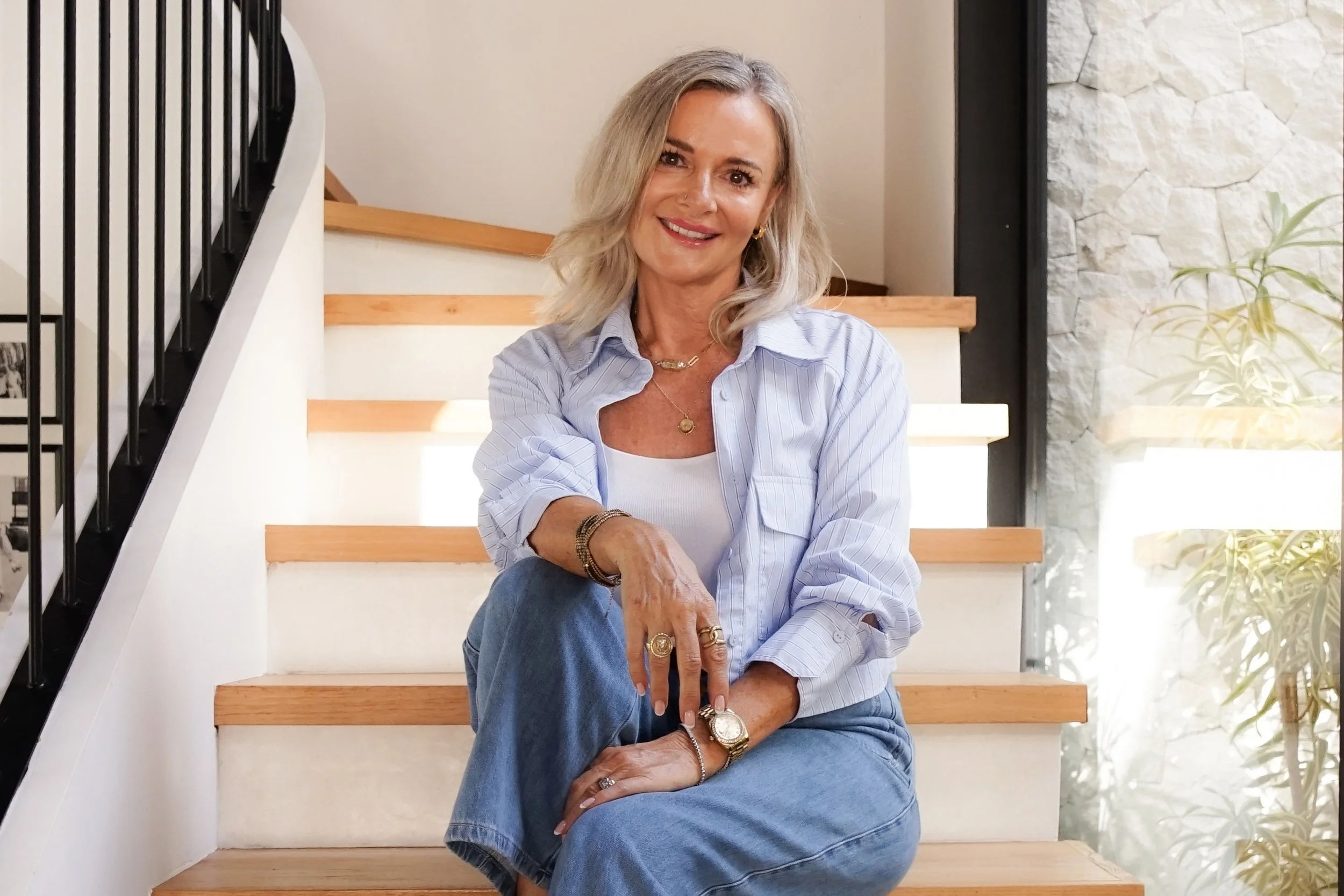 A woman sitting on wooden stairs with a railing, wearing a light blue striped shirt, white top, and blue jeans, smiling at the camera, with natural light coming from a large window on her right and plants visible outside.