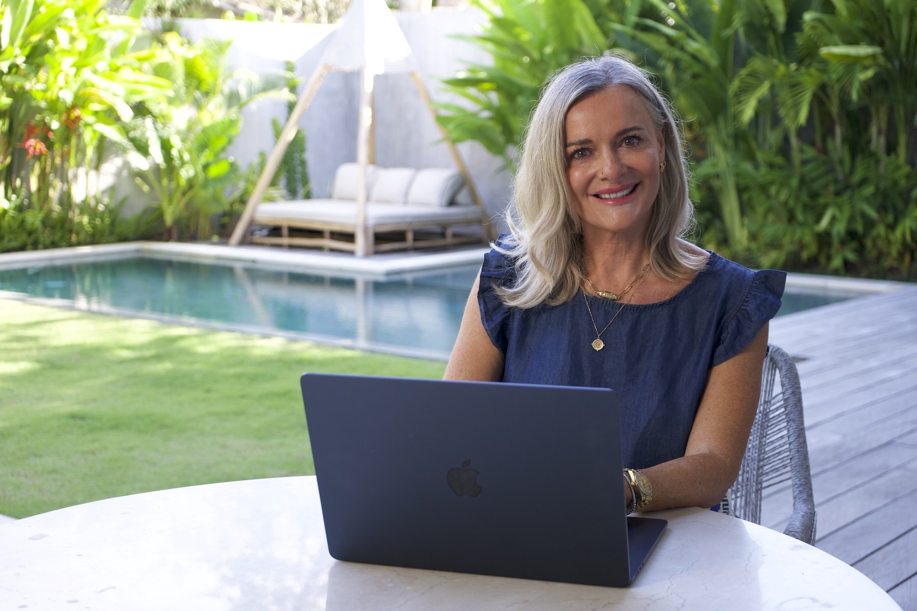 A smiling woman sitting at an outdoor table with a laptop in front of her, near a pool and lush greenery.