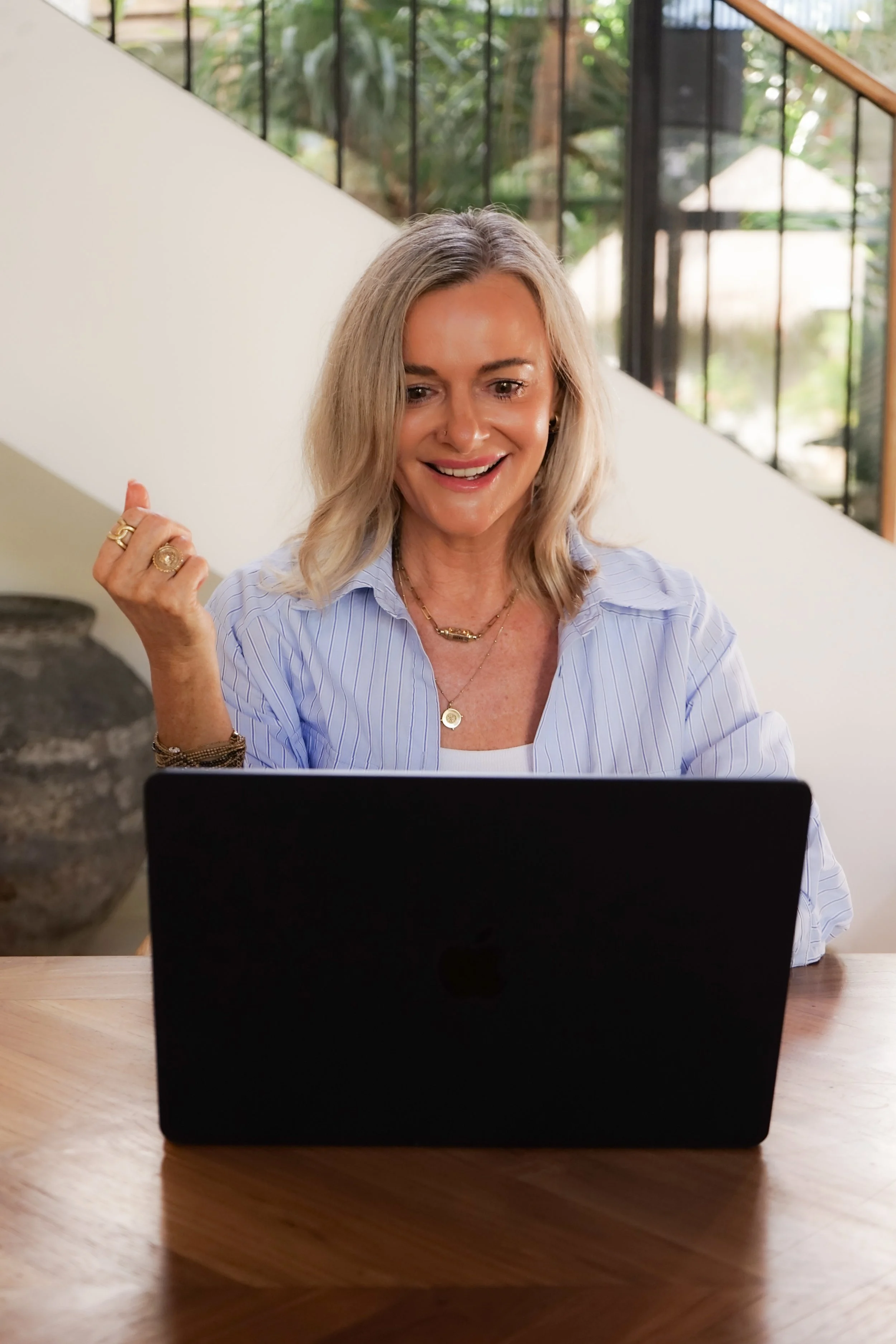A woman with blonde hair sitting at a wooden table, smiling, looking at a laptop, wearing a light blue shirt and jewelry, with a background of large windows and greenery.