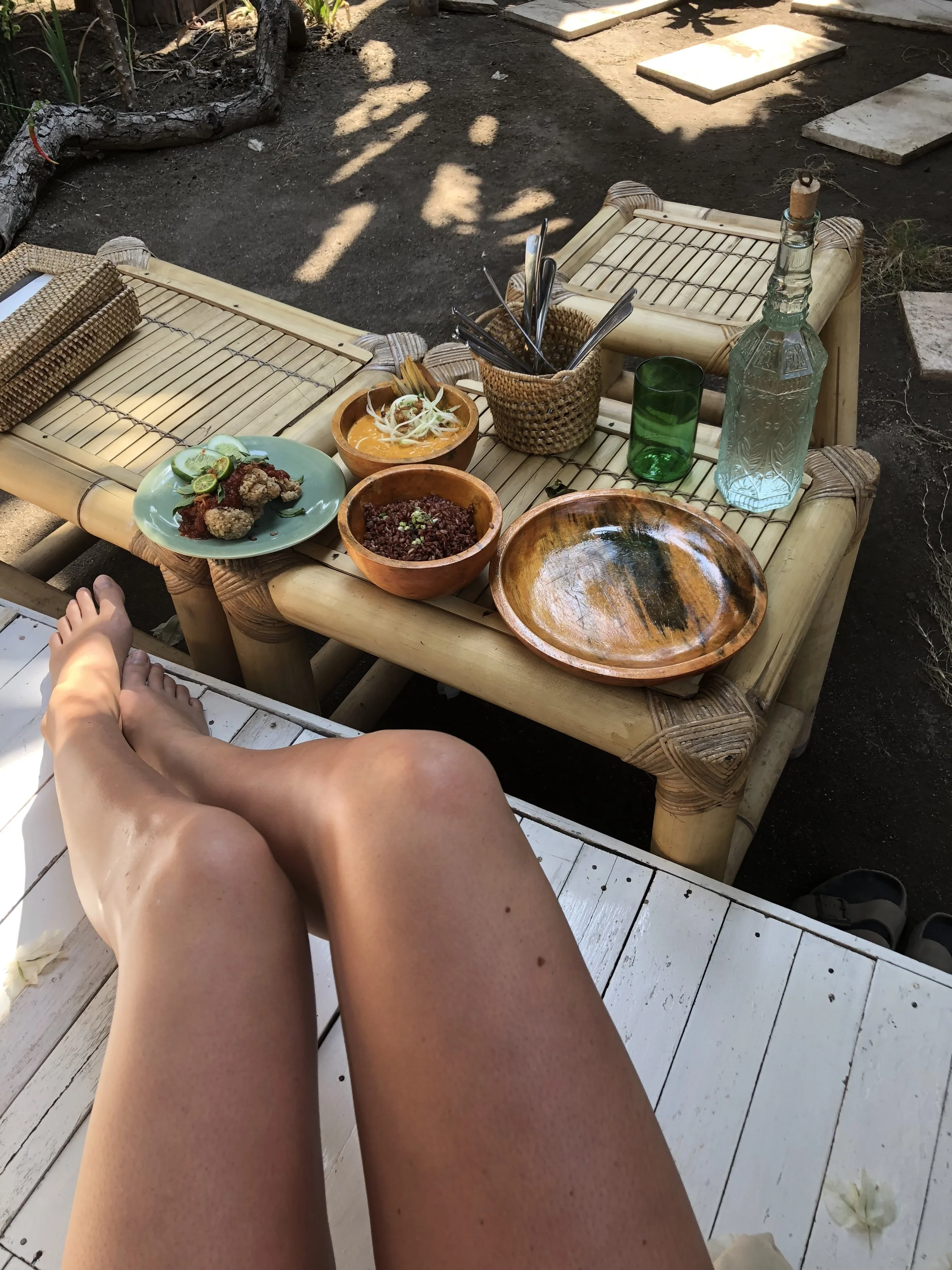 A person sitting outdoors with legs crossed, overlooking a bamboo table with plates of food, bowls, a green glass, a bottle, and utensils on it.