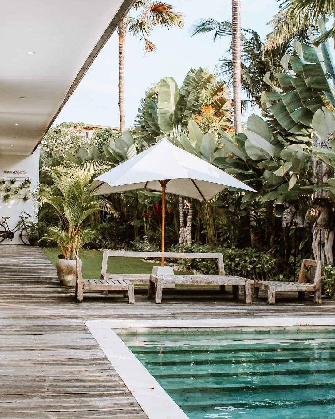 Poolside scene with wooden lounge chairs around a small swimming pool, a white patio umbrella, lush tropical plants, and tall palm trees under a clear blue sky.