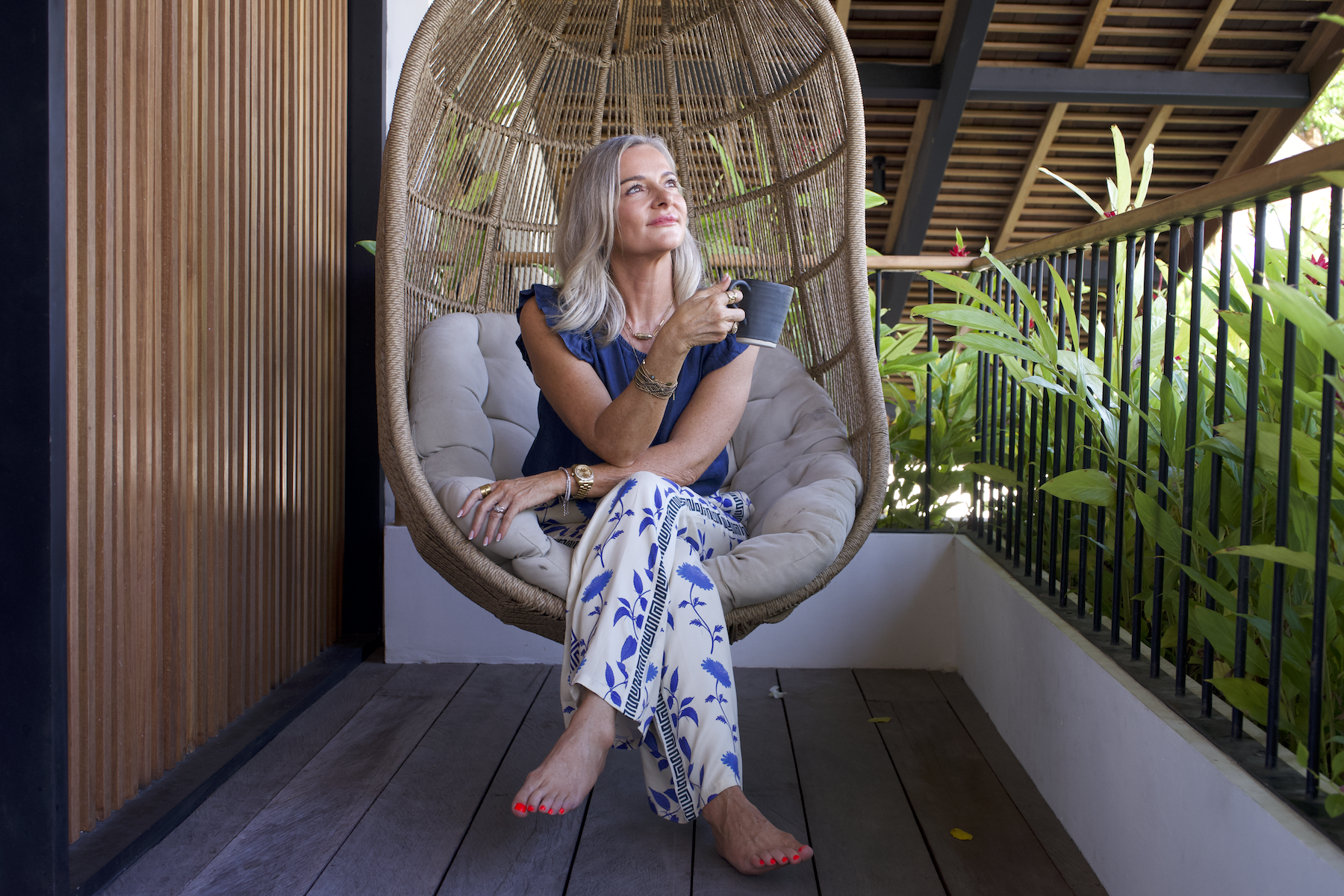 A woman wearing a navy blue top and patterned white and blue pants, sitting in a hanging wicker chair on a balcony, holding a black coffee mug, looking thoughtfully into the distance, with one foot resting on the floor and the other slightly raised.
