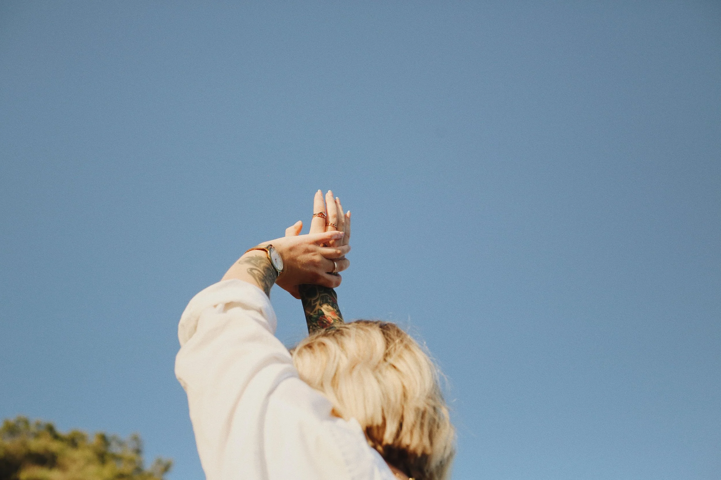 Person with tattoos and blonde hair stretching their arms upward against a clear blue sky.
