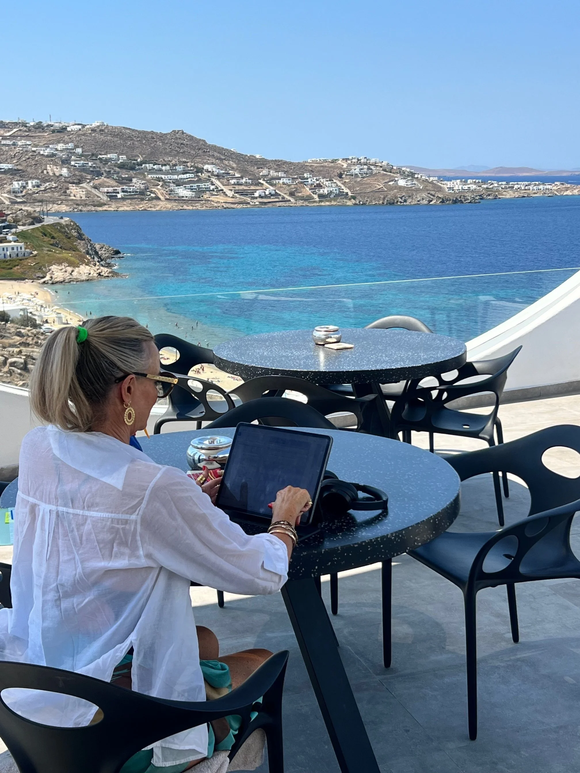 A woman sitting at a table on a terrace overlooking a blue ocean and distant islands, working on a tablet with a pair of headphones beside her.