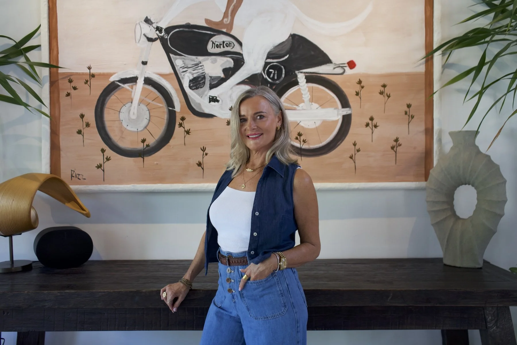 A woman with long blonde hair wearing a sleeveless denim shirt and high-waisted jeans, standing indoors in front of a large abstract painting of a motorcycle, with plants and decorative objects around her.