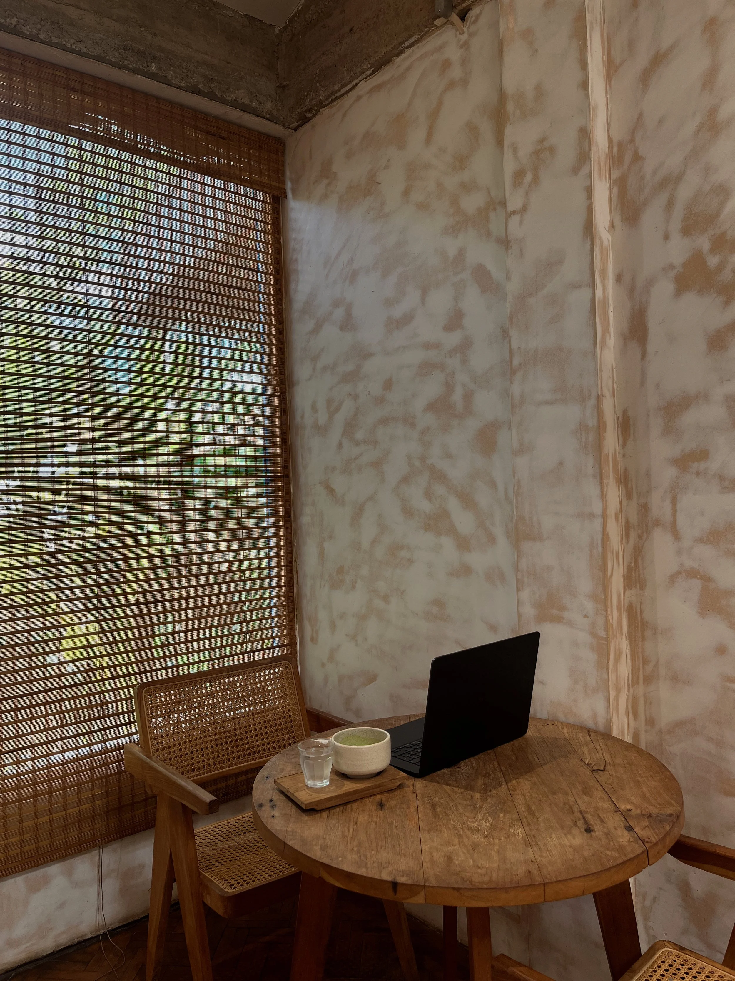 A cozy corner with a round wooden table, a bamboo chair, a laptop, a bowl of green tea, and a glass of water. The corner features a large window with bamboo blinds and textured walls.