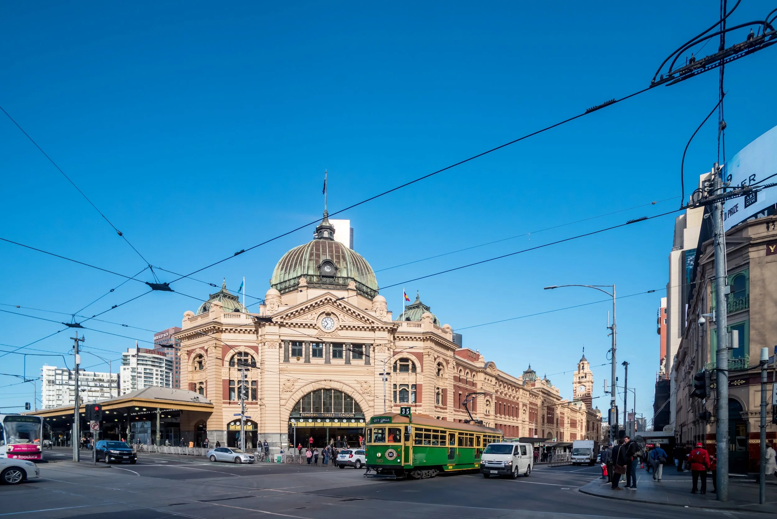 View of Flinders Street Station in Melbourne, Australia, with people and vehicles in front, a green tram, and overhead electric wires against a blue sky.