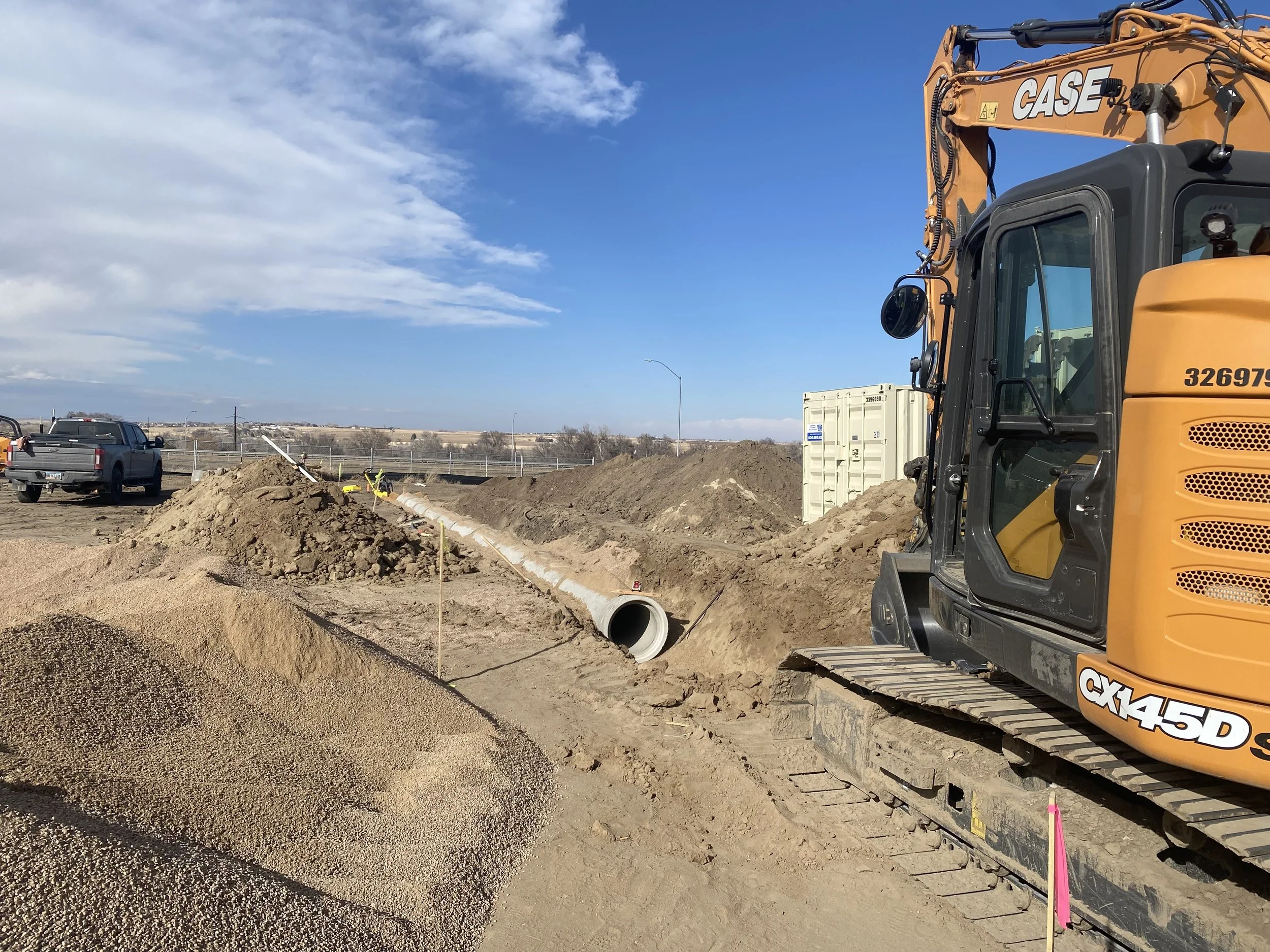 Construction site with a yellow CASE excavator digging a trench for a large concrete pipe, piles of dirt and gravel, a parked pickup truck, shipping containers, and partly cloudy sky.