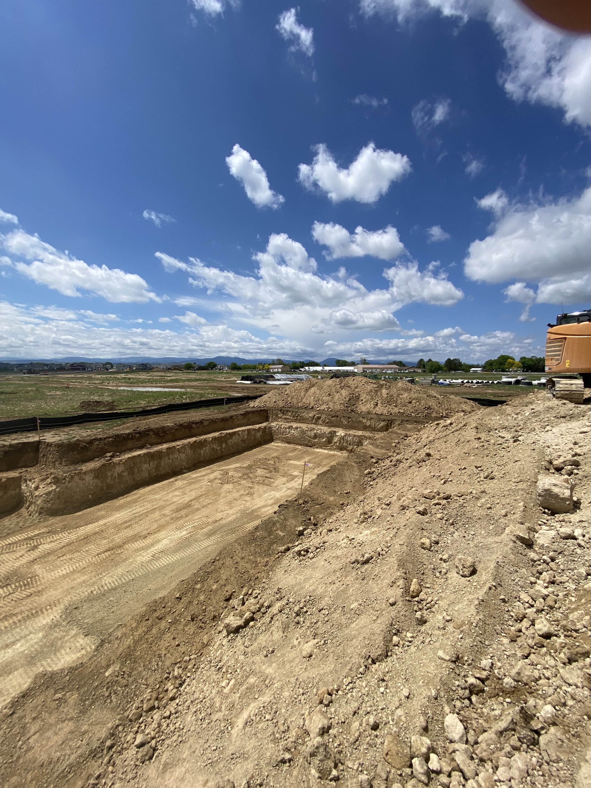 Construction site with excavated trenches, dirt piles, and a yellow excavator in the background under a partly cloudy sky.