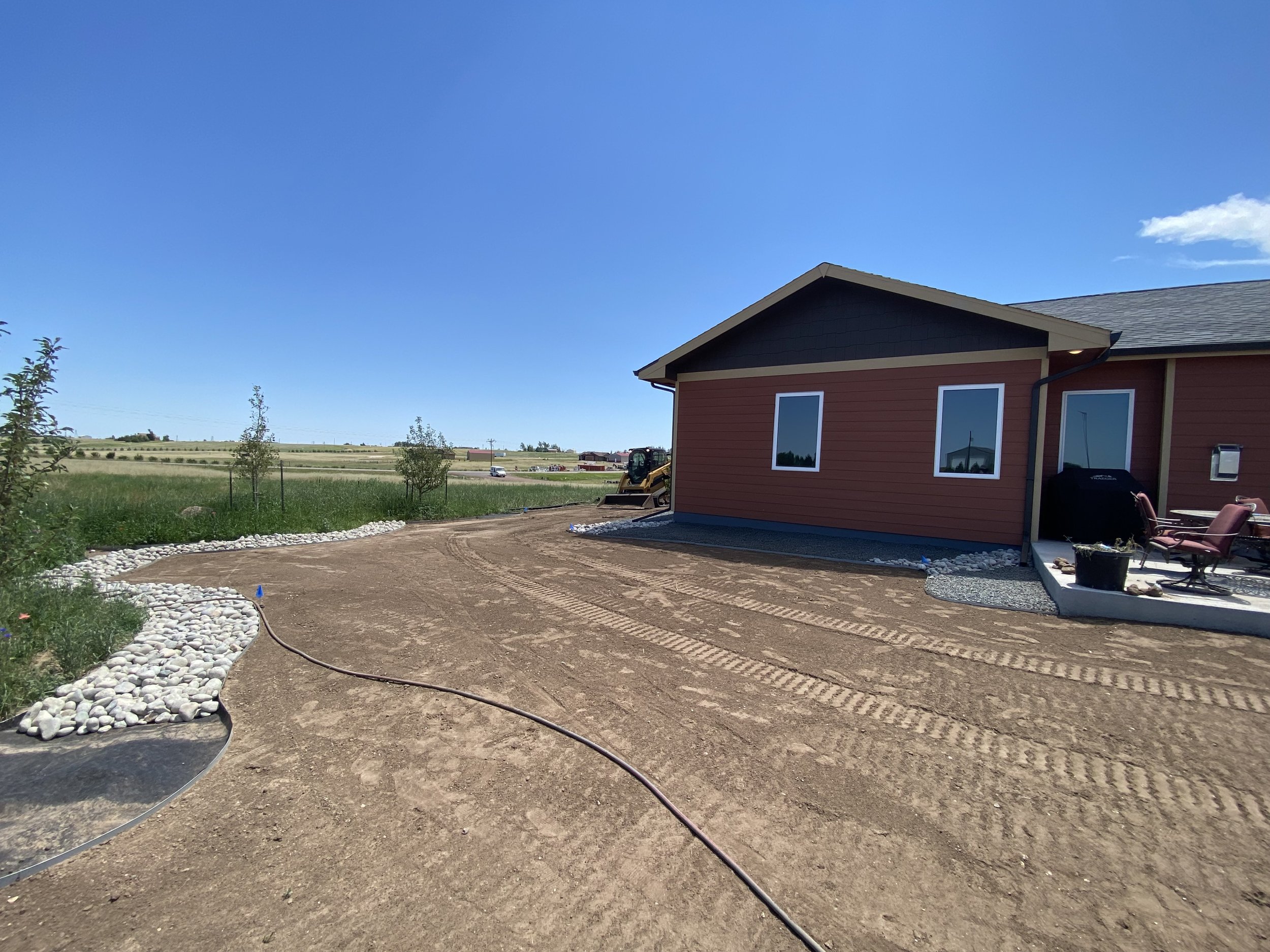 A newly landscaped backyard with dirt ground, white rocks bordering a garden bed, and a red house with patio furniture under a blue sky.