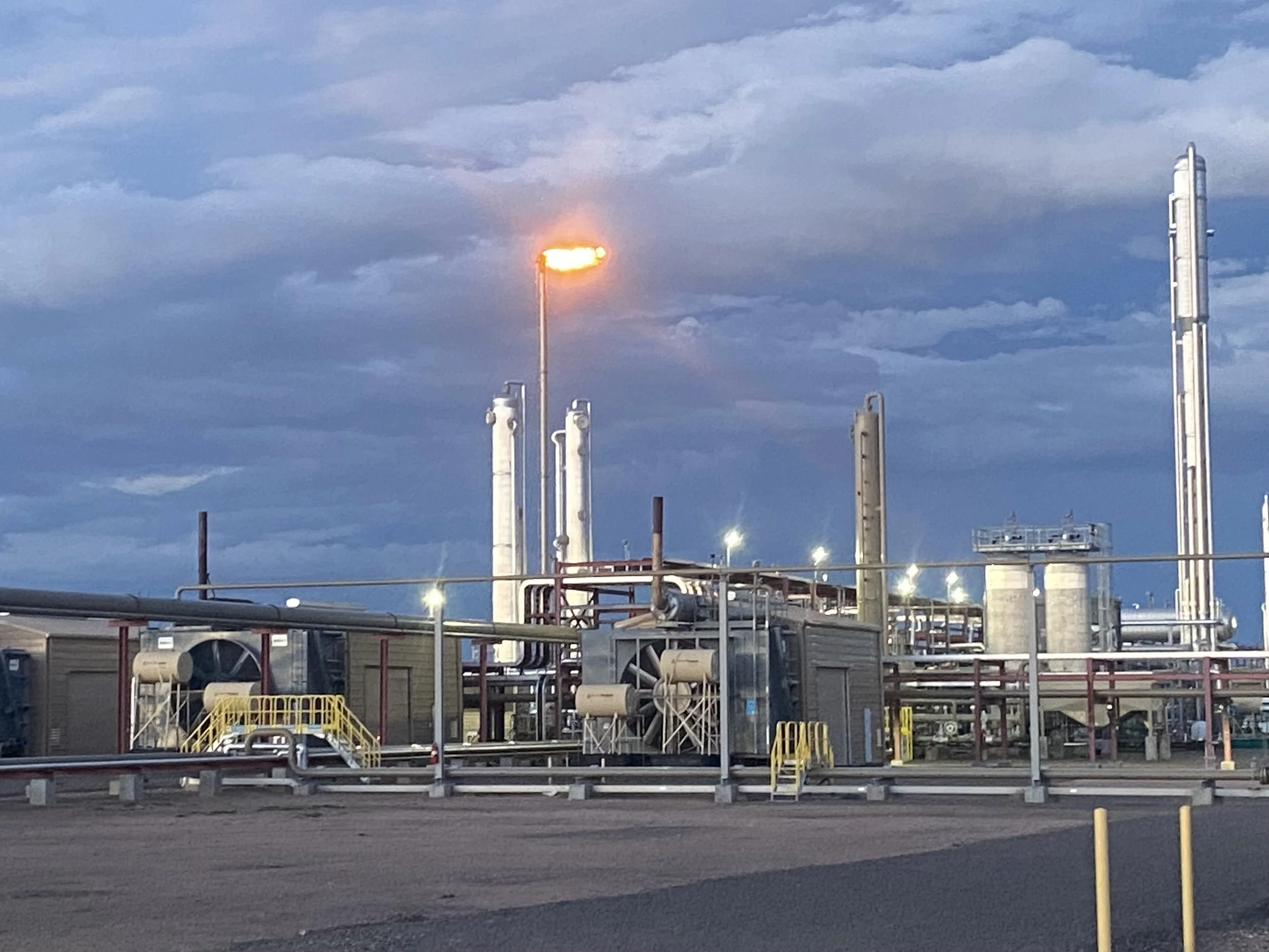An industrial oil refinery or chemical plant with tall distillation towers and equipment under a cloudy sky, illuminated by bright lights and a glowing flare.