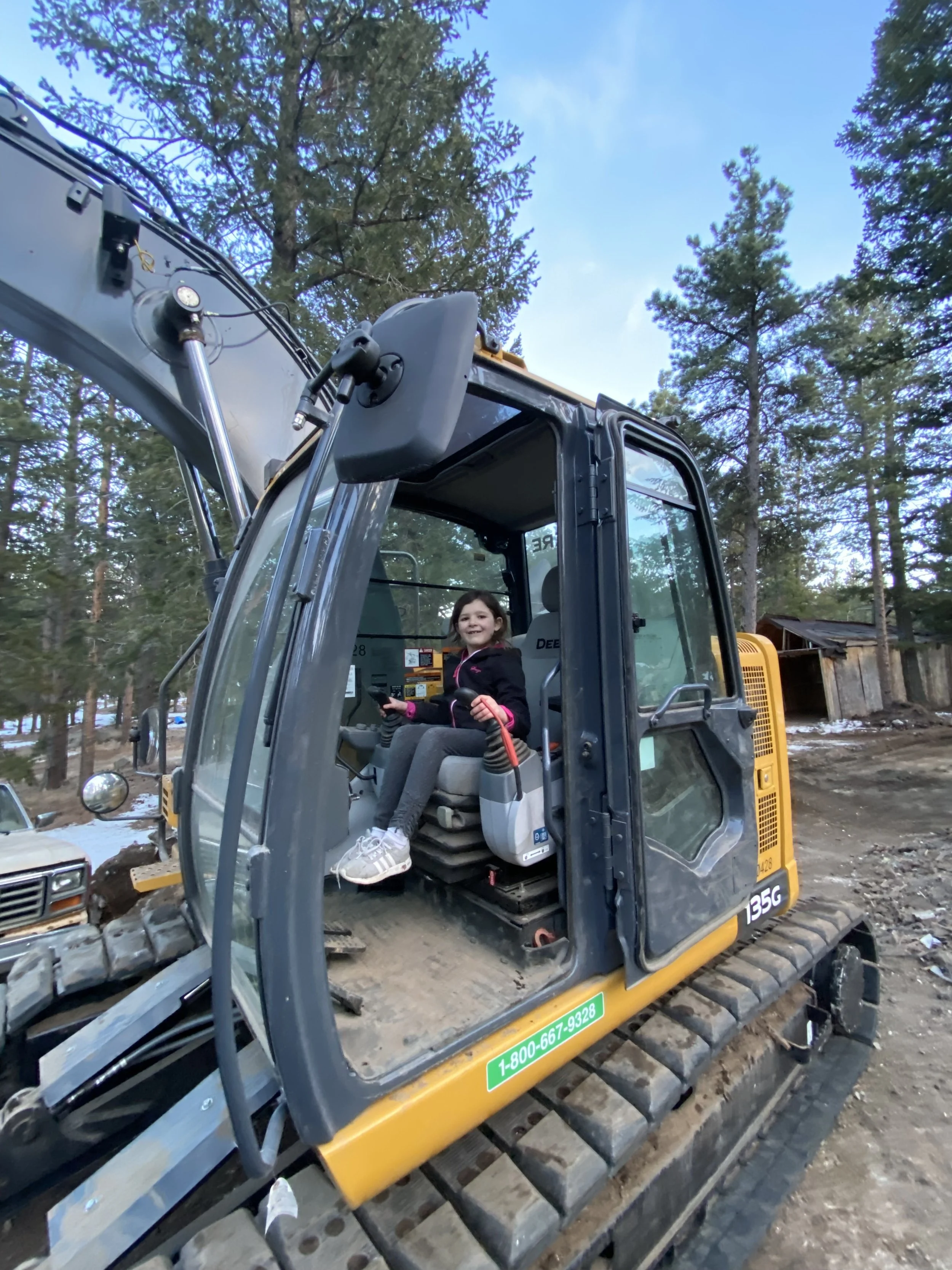 A young girl sitting inside the cab of construction equipment, with trees and a wooden shed in the background.